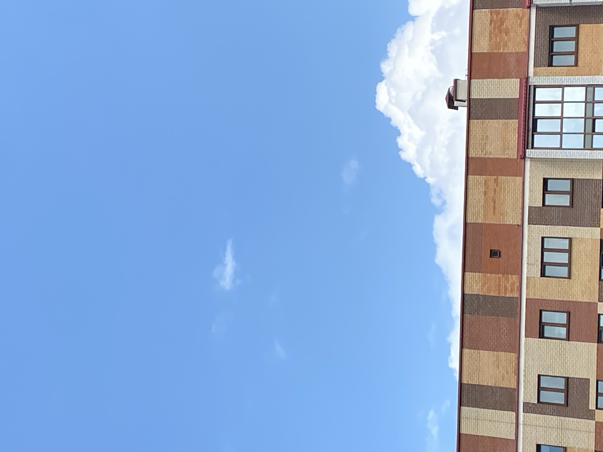 The image shows the upper portion of a modern apartment building against a bright blue sky with scattered clouds. The building features a facade constructed from bricks of varying shades of brown, red, and beige arranged in a patterned design. There are multiple windows visible, some rectangular and others with a grid pattern. A red roofline is visible along the top of the building, and a chimney with a conical cap protrudes from the roof. The scene appears to be an exterior shot, possibly taken during the daytime, given the bright sky. The architectural style suggests a contemporary urban setting.