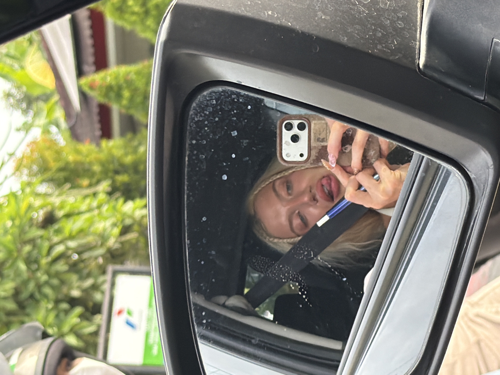 A blonde woman is reflected in a car's side mirror, sticking her tongue out and holding a blue pen. She is in the driver's seat of a car. The car is parked at a gas station in Mengwi, Indonesia. The gas station has green and white signage. The background shows trees and buildings.