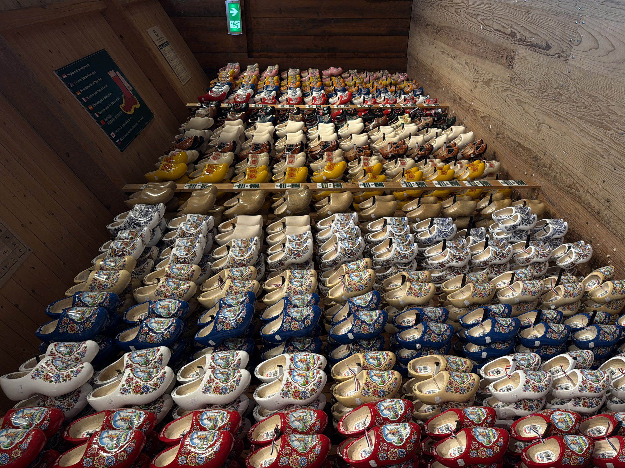 The image depicts a large collection of Dutch clogs, also known as klompen, displayed on wooden shelves against a wooden wall. The clogs are varied in color and design, with many featuring traditional Dutch motifs such as windmills and floral patterns. The collection is extensive, filling multiple shelves that extend along the wall. In the background, a wooden door is visible on the right side, and a green exit sign is mounted above it. The floor is made of distressed wooden planks. The overall impression is of a shop or museum dedicated to traditional Dutch footwear, likely located in a place like Zaandam, Netherlands, known for its historical windmills and cultural heritage.