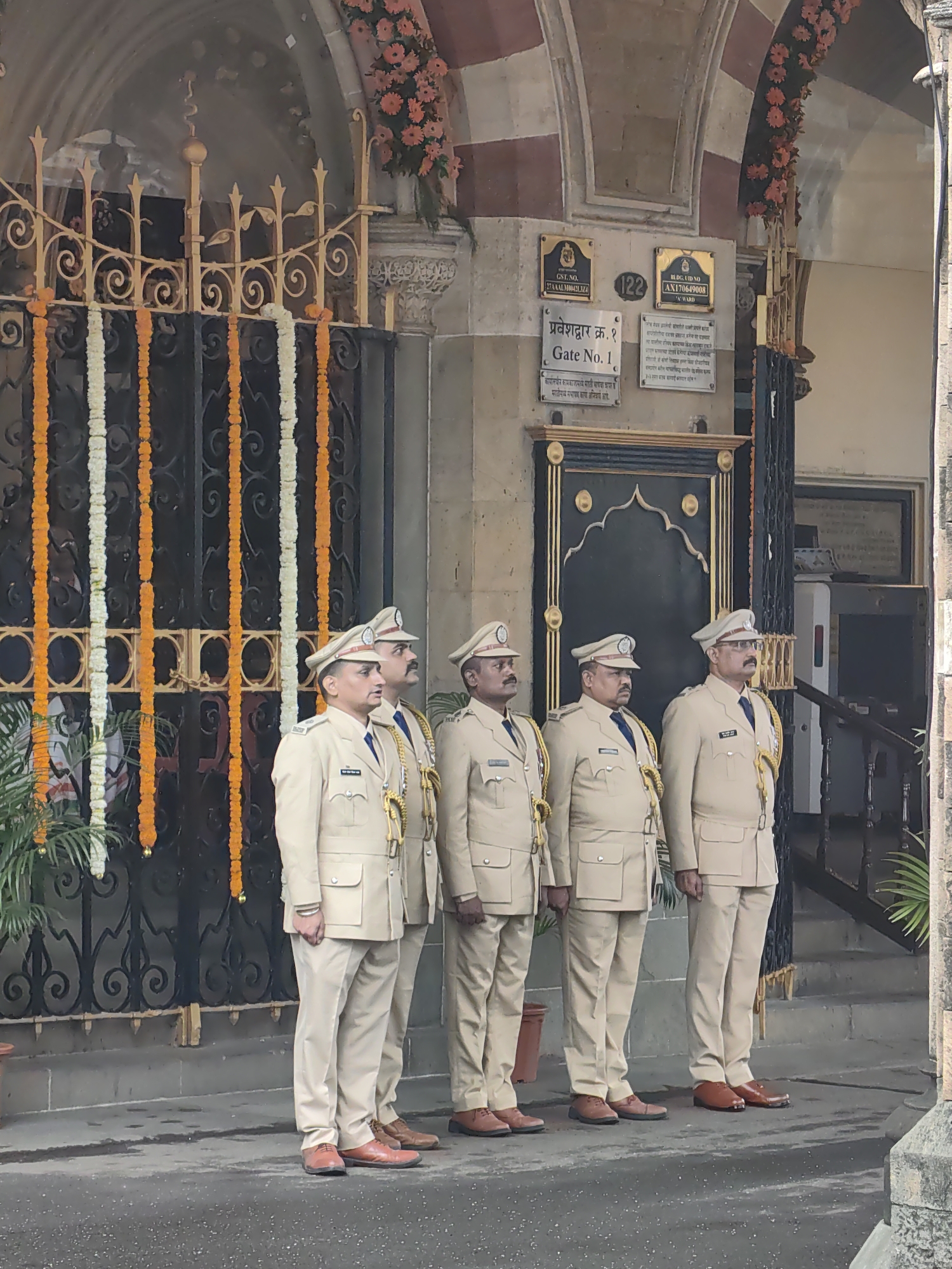 Five men in light-colored uniforms stand at attention in front of a dark metal gate decorated with garlands of orange and white flowers. The men are all wearing peaked caps and appear to be police officers. They are standing on a paved surface. Behind them, a building facade with ornate architectural details is visible. Signs are mounted on the wall, including one that reads "Gate No. 1" in English and a local script. Pink flowers are arranged in bunches on the building above the gate. The scene suggests a formal event or ceremony at an entrance to a building in Mumbai, India.
