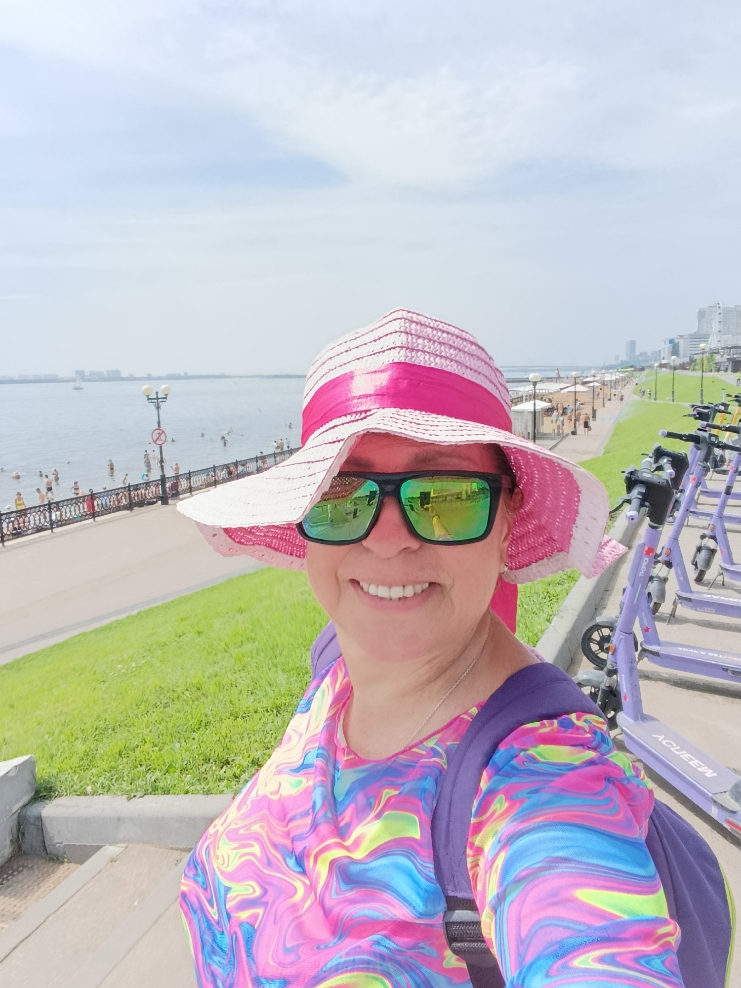 A woman, wearing a pink and white striped sun hat, large sunglasses with green lenses, and a brightly colored t-shirt with a swirling pattern, takes a selfie outdoors. Her reflection in the sunglasses shows a cityscape and a body of water. Behind her, a riverfront promenade is visible, with people walking and swimming in the water. Several purple electric scooters are parked in a line to her right. The sky is cloudy. The location is unknown.