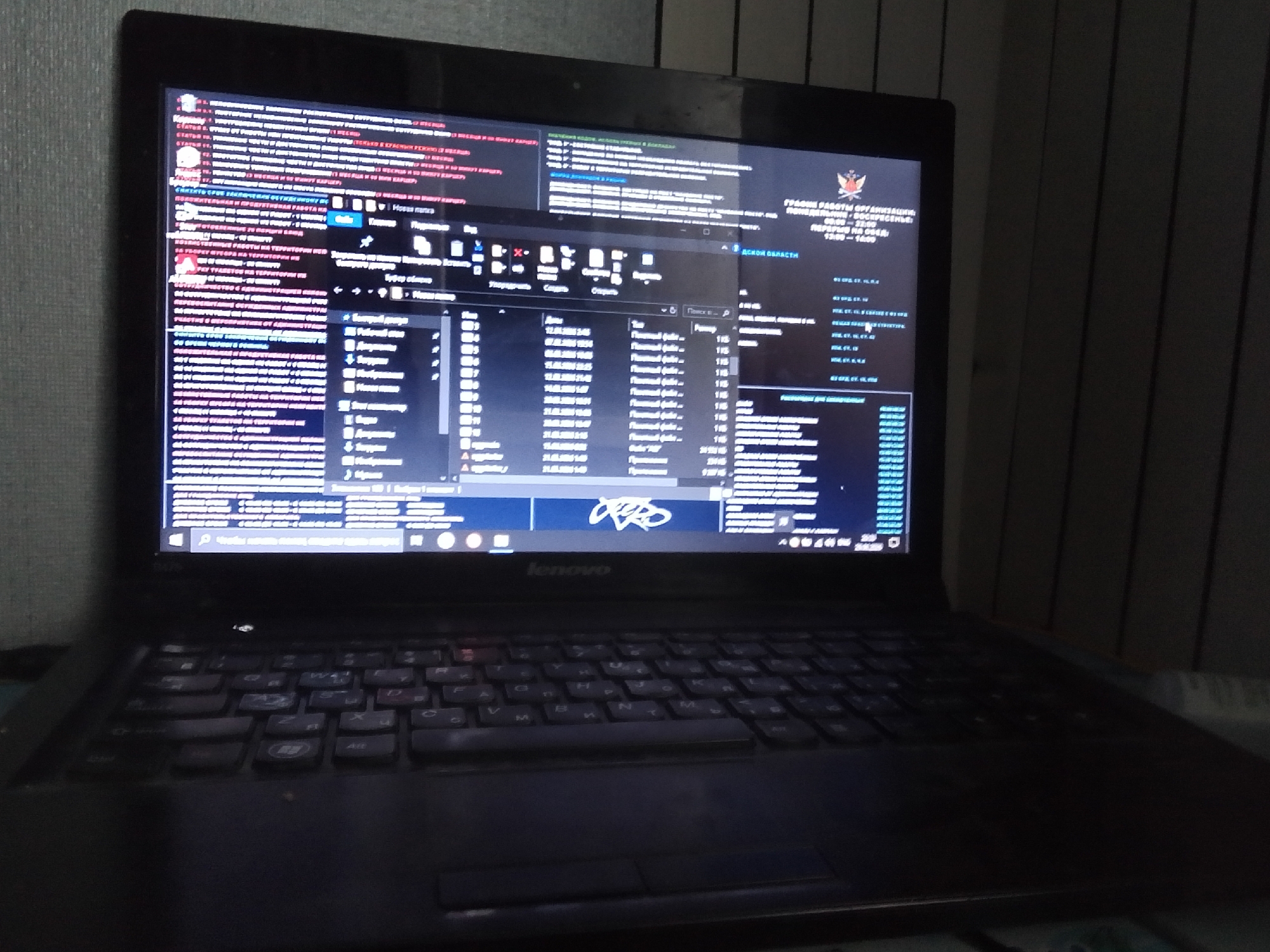 This is a close-up, dimly lit shot of a Lenovo laptop placed on a desk. The laptop screen is on, displaying a complex interface with multiple windows and a file explorer. The screen shows a mix of Russian text and what appears to be code or system information.
The primary subject is the laptop itself, along with its illuminated screen. The screen displays what looks like a work-related application or system status, with various panes and lists of data. One prominent window is a file explorer, showing file names, dates, and sizes. There's also a graphic resembling a logo on the screen, possibly for an organization.
The scene is dark, suggesting it's either nighttime or the room is otherwise dimly lit. The background consists of vertical blinds or shutters, indicating an indoor setting. There are no people visible in the frame.
Visible text on the screen includes: "ГЛАВНЫЕ РАБОТЫ ОРГАНИЗАЦИИ", "ПЕРЕРЫВ НА ОБЕД", "13:00 - 14:00", "ОСНОВНАЯ ОБЛАСТЬ", and numerous other lines of text in Russian, some of which appear to be file paths or data entries. The file explorer window has column headers like "Имя", "Дата изменения", and "Тип". The brand name "Lenovo" is visible on the laptop's bezel below the screen.
The overall impression is one of focused work or activity on the computer in a private, perhaps office or home, environment. The low light and the content on the screen hint at a serious or professional task being undertaken. The location context of Kalachinsk, Russia, suggests the text on the screen is likely in Russian.