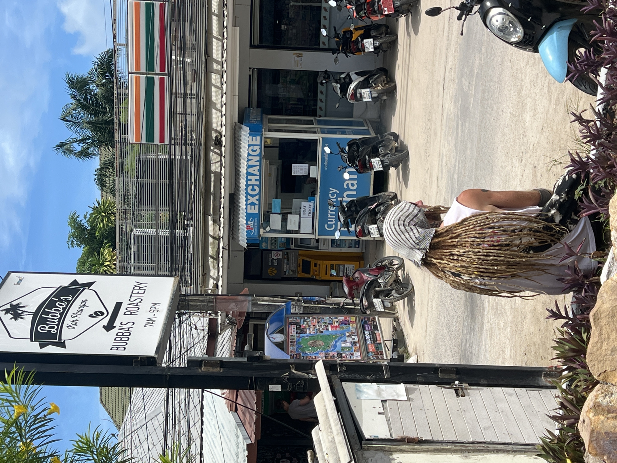 In the town of Tambon Koh Phangan, Thailand, a street scene unfolds under a bright, clear blue sky with scattered clouds. Dominating the left side of the frame is a black and white sign for "Bubba's Koh Phangan" and "Bubba's Roastery," indicating operating hours of 7 AM to 5 PM. To the right of the sign, a row of buildings with corrugated roofs lines the street. These buildings house businesses, including a currency exchange booth with a blue and white striped awning displaying "PM EXCHANGE" and "Currency Exchange." Next to the exchange booth, a 7-Eleven convenience store is visible, identified by its iconic red, green, and orange stripes on the upper facade.
Parked along the street are numerous scooters and motorcycles, suggesting a common mode of transportation in the area. In the foreground, a person with long dreadlocks, wearing a white tank top and a striped head covering, is seated with their back to the camera. They are positioned in front of a light-colored wooden structure with a metal latch on its door. In the mid-ground, a red motorcycle is parked near a display board featuring a map and various advertisements. A man in a grey t-shirt is walking away from the viewer near the entrance of one of the buildings on the left. The environment appears to be a commercial area with overhead electrical wires strung across the scene.