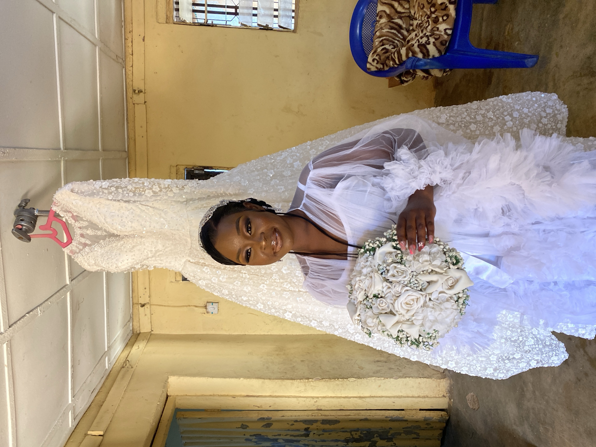 A woman in a white wedding dress and veil is holding a bouquet of white flowers and smiling at the camera. The dress is heavily embellished. A white wedding dress is hanging in the background on a pink hanger. A blue chair with animal print fabric is in the background. The photo appears to be taken indoors in a room with yellow walls, with Vom, Nigeria as the location.