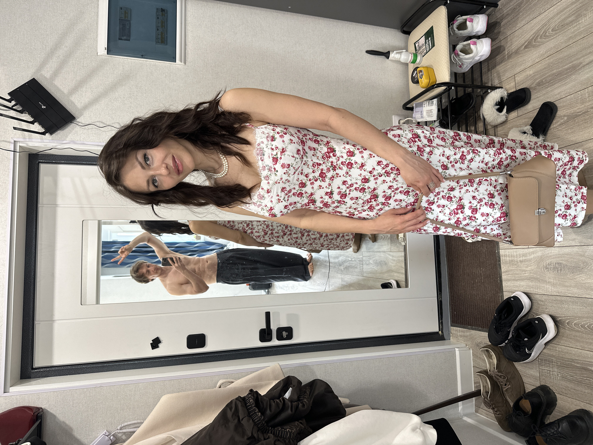 The image shows a woman standing in front of a mirror, apparently in an apartment in Murino, Russia. The woman is wearing a floral dress and a pearl necklace. She is holding a small purse. In the reflection of the mirror, a shirtless man is taking a selfie. In the background, there is a router on the wall, a breaker box, a shoe rack with shoes on it, and some other clothing items. The apartment has light-colored walls and wood-patterned flooring. There are also shoes on the floor near the mirror.