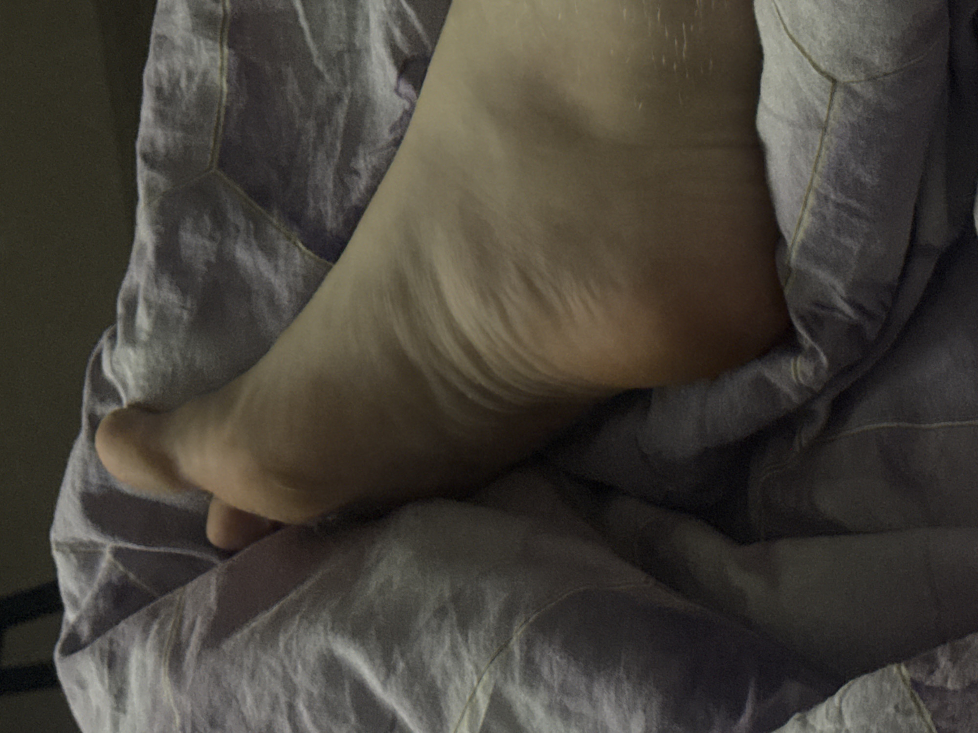 The image shows a close-up of a person's bare foot and ankle, partially covered by a lavender-colored blanket with a hexagonal pattern. The foot is positioned on its side, with the toes facing left and the heel to the right. The skin appears smooth with some visible creases around the ankle and the sole of the foot. The lighting is dim, casting soft shadows and highlighting the contours of the foot. The blanket is soft and wrinkled, suggesting a comfortable or relaxed setting. There is no indication of the location being in Saratov, Russia, or any specific urban environment. The scene appears to be an indoor setting, possibly a bedroom.
