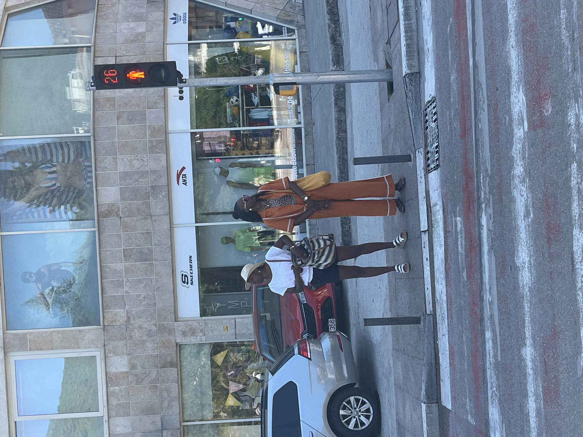 This image captures an urban street scene in Budva, Montenegro, under bright daylight. In the foreground, a faded pedestrian crossing with white and red stripes is visible on the asphalt road. A concrete sidewalk lines the road, featuring two dark bollards and a drainage grate near the curb.
Two adult females are positioned on the sidewalk near a pedestrian traffic light. This light, mounted on a pole to the right, displays a red standing figure and the countdown "26". The female on the left wears a white t-shirt, dark shorts, a light straw hat, and light sandals, and is holding a striped shoulder bag while looking downwards. The female on the right wears an orange two-piece outfit with white trim, a patterned top underneath, sunglasses, and flat sandals, and carries a yellow shoulder bag while looking to her left.
To the left of the pedestrians, a silver Volkswagen Golf is partially visible, followed by a red Renault Clio with a license plate displaying "356". The background is dominated by a multi-story building featuring a facade of light-colored square tiles. The ground floor houses several retail stores, identifiable by their large glass windows and signage for "SKECHERS", "ANTA", and "adidas". Upper-story windows reflect a green hillside and display large-scale images, including a man on a surfboard and a woman wearing a striped bikini top and an open striped shirt.