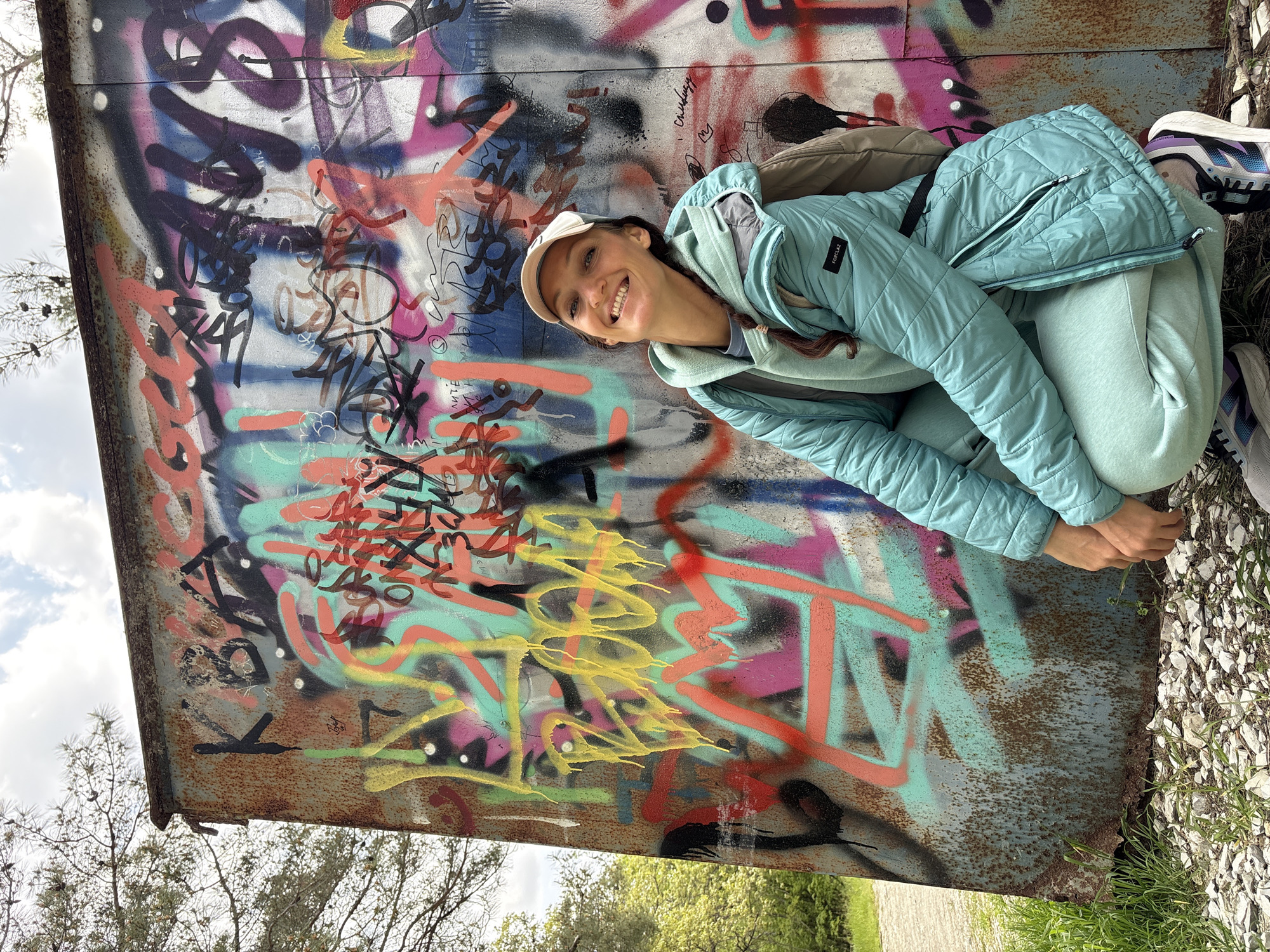 A woman is crouching next to a wall with graffiti on it. The wall is made of rusted metal. The graffiti is in a variety of colors, including black, red, yellow, pink, purple, and teal. The woman is wearing a light blue quilted jacket, matching sweatpants, and a cream-colored baseball cap. She is smiling and looking at the camera. A tan backpack is on her shoulders. Some of her athletic shoes are visible to the right side of the image. A patch of green vegetation is on the ground next to the right side of the wall. Some trees are in the background and the corner of the sky is visible in the upper left corner. The location is Gelendzhik, Russia.