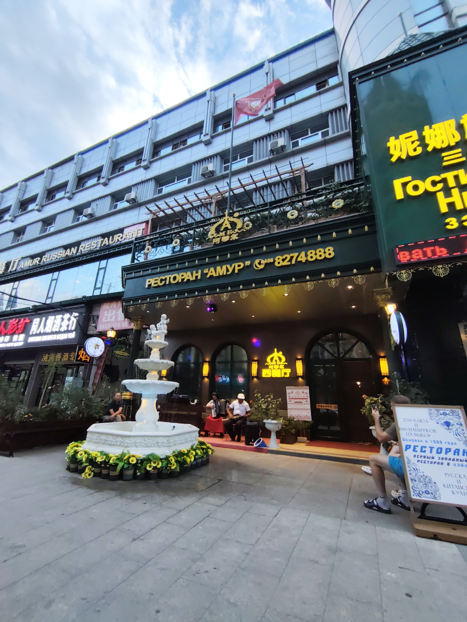 The image captures a street scene in Aihui, China, featuring a restaurant with a prominent Russian theme. The restaurant, named "RESTORAN AMUR", is identifiable by its Cyrillic lettering and a phone number displayed. A multi-tiered white fountain with surrounding sunflowers sits in front of the establishment. Two musicians, one playing an accordion and the other a stringed instrument, are visible performing near the fountain. Several people are present in the scene, some seated and others standing. The architecture suggests a commercial area with shops and businesses lining the street. The sky above is partly cloudy.