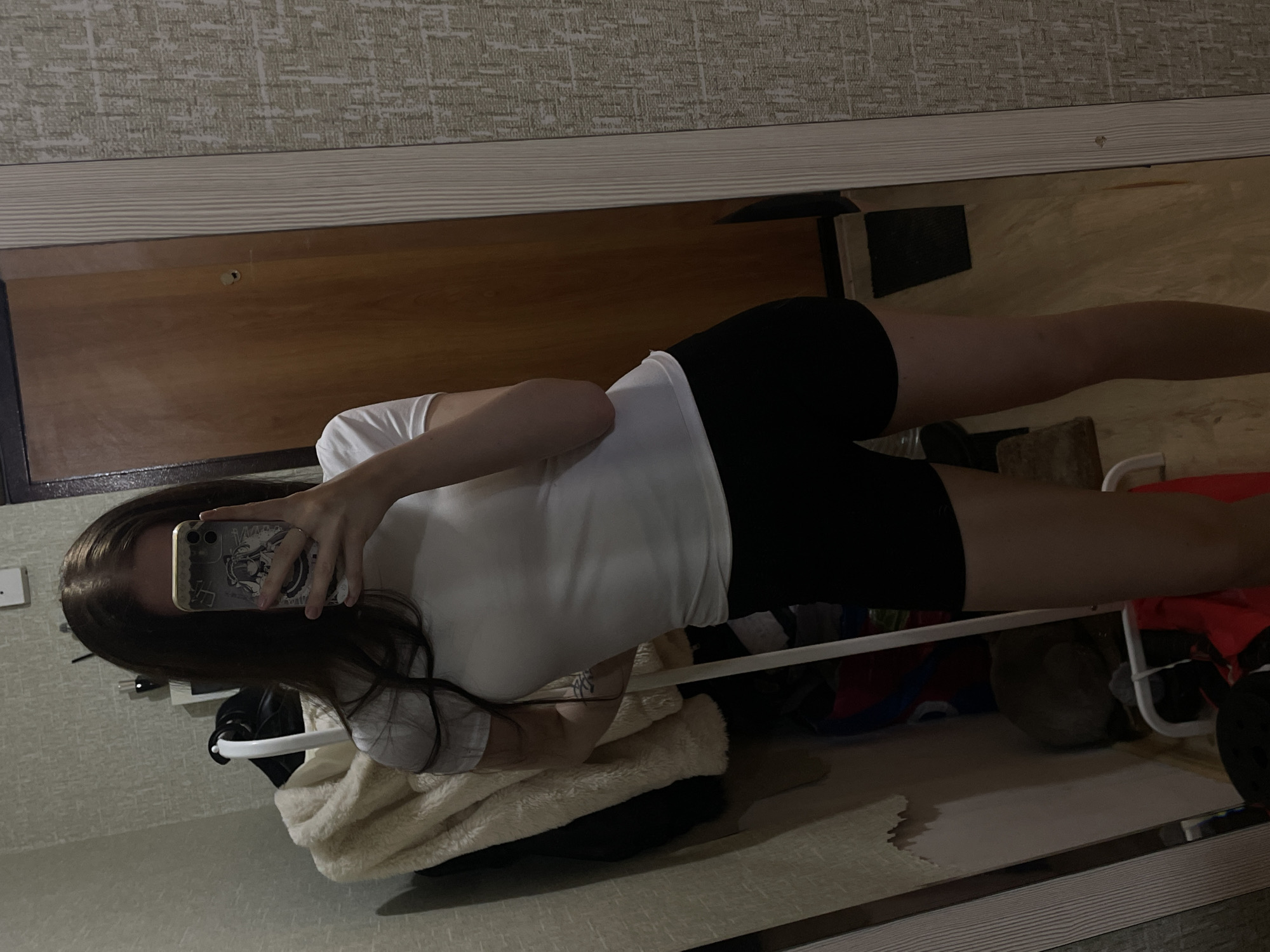 The image depicts an indoor scene in the city of Votkinsk, Russia. A person, appearing to be female, with long brown hair, is captured in a full-length mirror selfie. Her face is largely obscured by a gold-colored smartphone, held in her right hand, which features a distinctive white and purple anime-themed case and a dual-lens camera system. She is wearing a form-fitting white short-sleeved t-shirt and plain black shorts.
The mirror, framed by a light-colored, vertically ridged border on the right, reflects a section of a textured, light-grey wallpaper with an irregular pattern on the right side of the scene. To the right in the reflection, a light-to-medium brown wooden door is visible, featuring a circular peephole and a small, dark rectangular mat on the light wooden floor directly in front of it.
Behind the person, to the left, is another section of the same textured wallpaper, with a small area of peeling wallpaper near the bottom left, revealing a lighter surface beneath. A thick, fluffy, cream-colored coat or jacket hangs on a white garment rack or stand directly behind the person, covering other darker garments and various items, including blue and red bags, resting on the floor. A white, utilitarian-style rack or stand is also partially visible at the bottom left. In the upper left background, a white rectangular wall fixture, possibly a light switch or thermostat, is mounted. The overall lighting appears dim, suggesting an indoor setting with limited natural light or subdued artificial illumination.