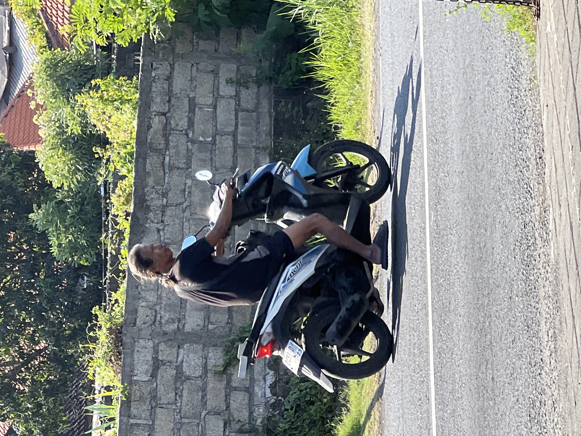 A Balinese man with long hair pulled back in a ponytail rides a blue and white scooter down a paved road in Denpasar, Indonesia. He is wearing a black t-shirt and dark shorts, and appears to be wearing flip-flops. The scooter has a license plate that reads "2431 DJ". Behind the man, a low stone wall runs along the side of the road, topped with lush green foliage and glimpses of red-tiled roofs. The sun is shining brightly, casting shadows on the road, indicating it is likely daytime. The background is filled with trees and vegetation, suggesting a tropical environment.