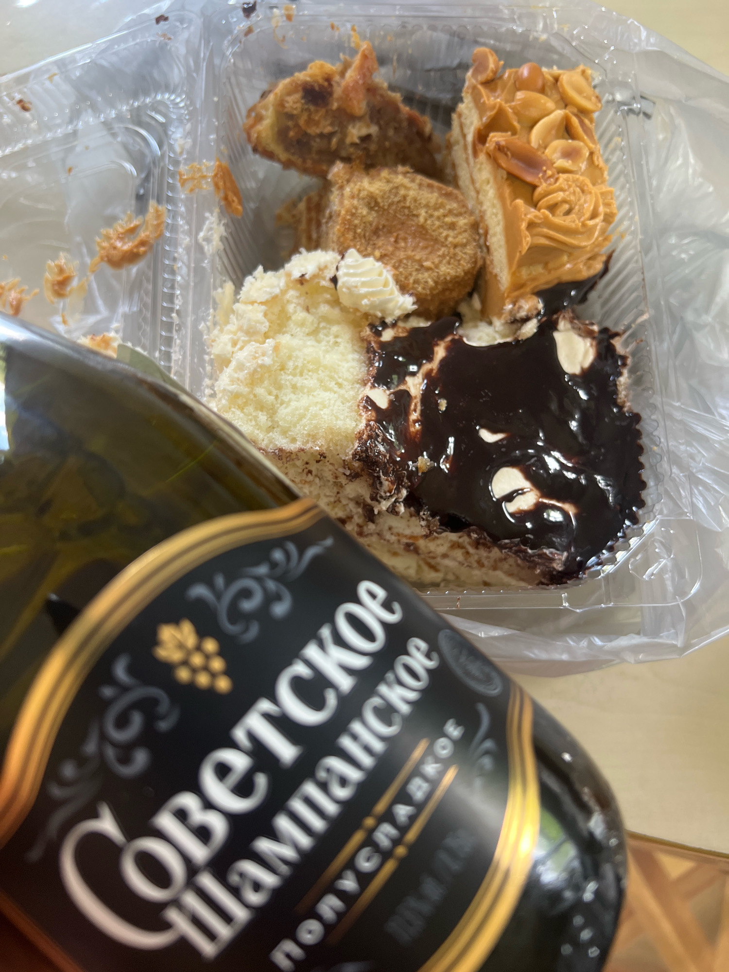 A bottle of Soviet Champagne with Cyrillic lettering is held in the foreground, obscuring a partially eaten assortment of pastries in a clear plastic container. The pastries appear to be a slice of cake with chocolate frosting, a piece topped with caramel and peanuts, and a smaller, round pastry. Crumbs and frosting are visible in the container and on its lid. The background is blurred, but appears to be a wooden table surface. The scene is likely indoors, as suggested by the lighting and reflections on the bottle. There are no people visible in the image. The location of Sokhumi, Abkhazia is not indicated in the image itself.