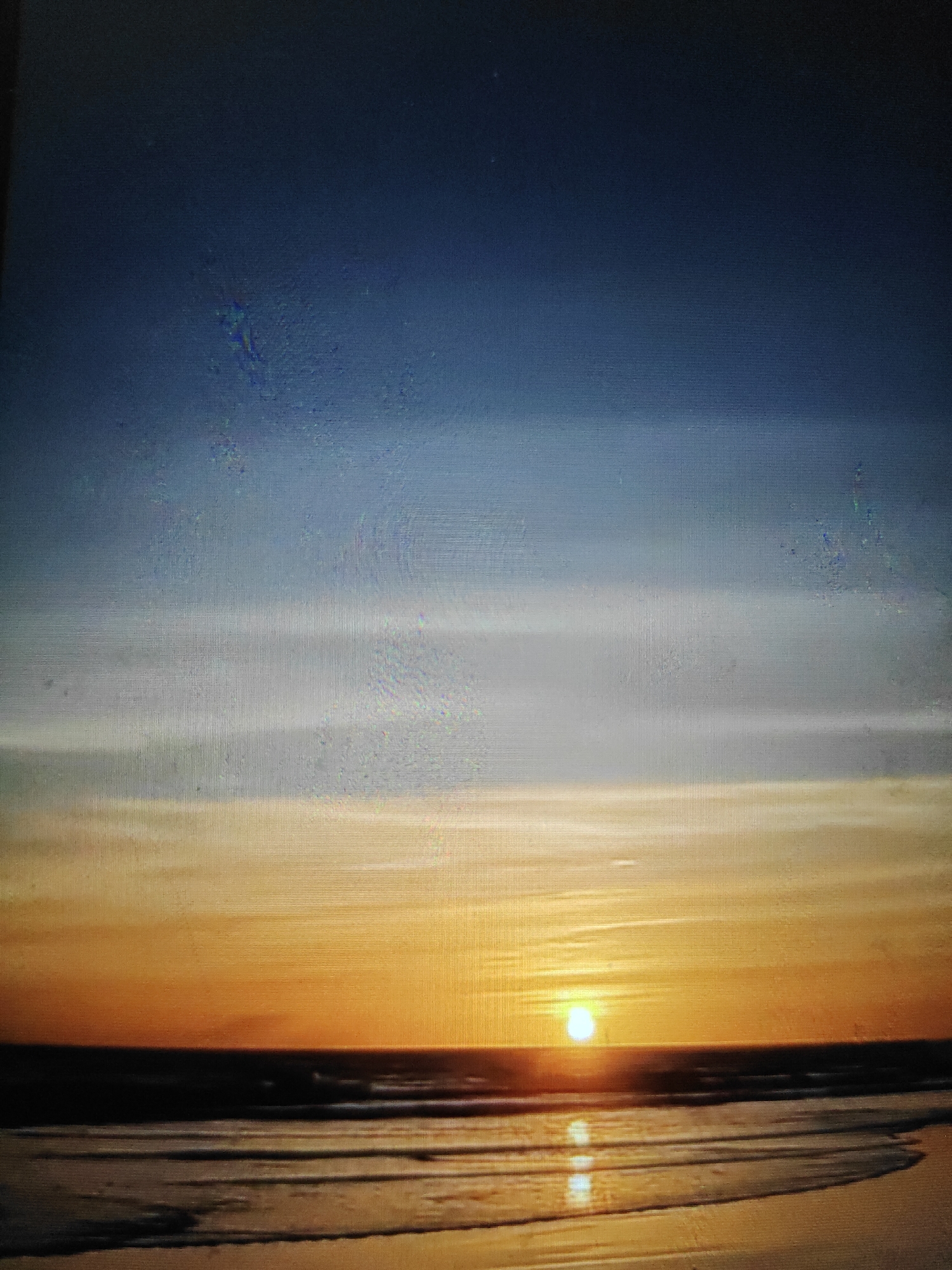 The image depicts a sunset over the ocean at a beach. The sky transitions from a deep blue at the top to lighter shades of blue and white, then to vibrant orange and yellow hues near the horizon. A bright sun is visible low in the sky, casting a reflection on the wet sand and the gentle waves that are breaking on the shore. The water appears dark, contrasting with the illuminated sky and sand. There are no people or specific objects clearly visible in the scene, other than the natural elements of sky, sun, ocean, and beach. The environment is coastal and peaceful, indicative of a sunset by the sea.