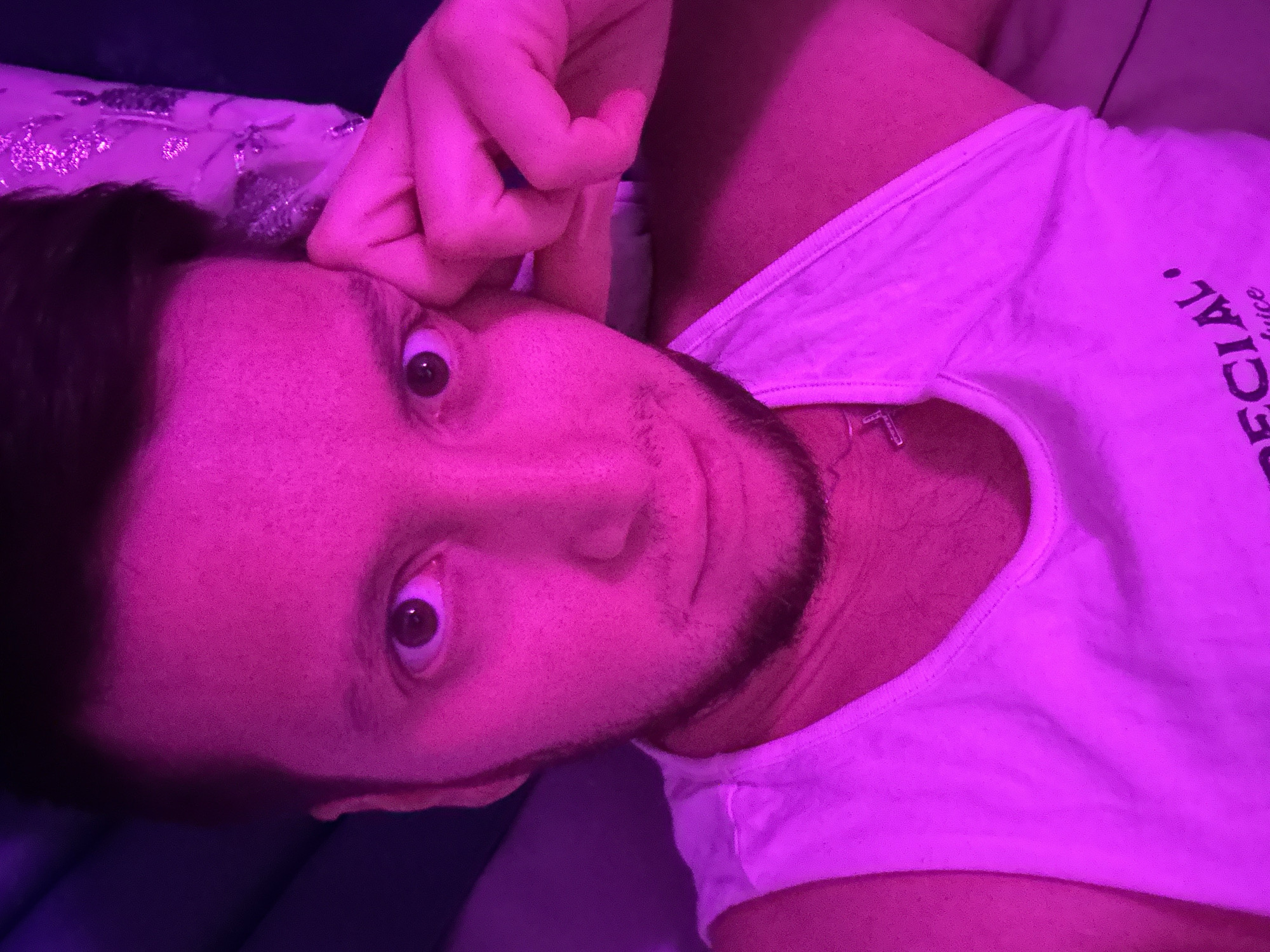 The image is a close-up, high-angle selfie of a fair-skinned man with dark hair and a short beard, lit by pink and purple light. He is wearing a white tank top with black text on the front, partially visible. His left hand is raised and resting against his face, near his eye and temple. His eyes are open and looking at the camera. The background is dark and out of focus, with some textured purple fabric visible on the right side, possibly a pillow or blanket. The scene appears to be indoors, possibly a bedroom. No specific landmarks or details indicate a location in Mirny, Russia.