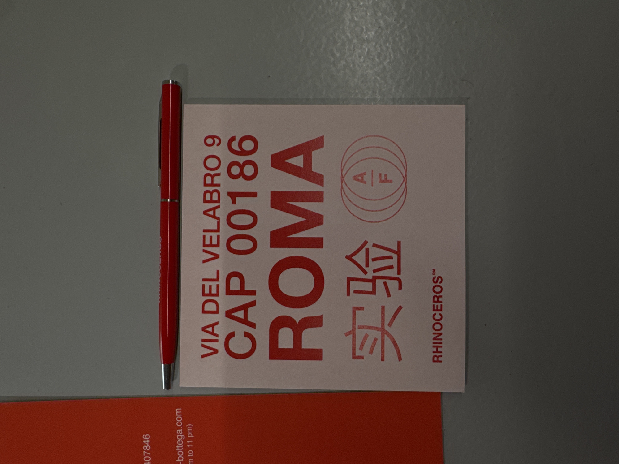 A red pen lies next to a light pink card that contains a postal address and a brand name. The text on the card in red ink reads “VIA DEL VELABRO 9 CAP 00186 ROMA”. Below the address is the Chinese characters for experiment (实验) in red ink. Below that is the brand name RHINOCEROS™ in red ink. A partially visible orange card is at the bottom of the image. The text on that card reads “407846” on one line and “-bottega.com m to 11 pm)” on the next line. The location in Rome is Via del Velabro.