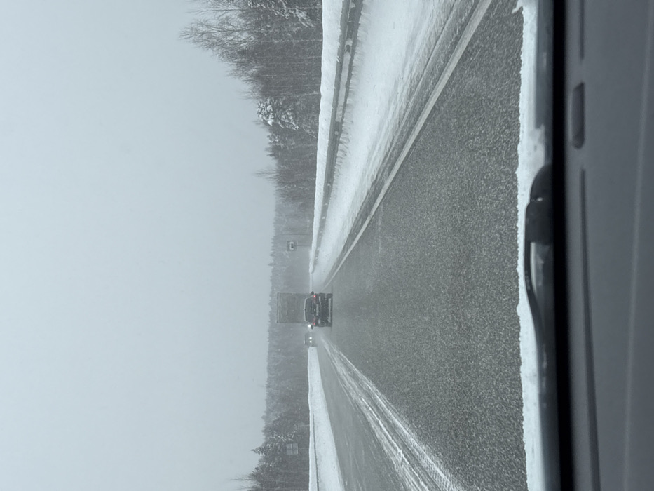 The image depicts a snowy highway scene during what appears to be a winter storm or heavy snowfall. The perspective is from inside a vehicle, looking straight ahead through the windshield.
The road is a multi-lane asphalt highway, partially covered with a thin layer of snow or slush. Tire tracks are visible in the snow on the right side of the road, suggesting recent vehicle passage. To the right of the asphalt lanes, there's a snow-covered shoulder or median barrier, which has a significant accumulation of snow and ice, creating an uneven, textured surface.
In the distance, two vehicles are visible traveling in the same direction as the viewer. The foremost vehicle is a large, dark-colored truck, possibly a semi-trailer or box truck. Behind the truck, a smaller passenger vehicle, dark in color, can be seen with its red taillights faintly glowing, indicating its brake lights or running lights are on.
To the far right of the road, beyond the snow-covered barrier, is a dense forest of trees, heavily laden with snow. The trees appear to be a mix of evergreens and bare deciduous trees, all contributing to the wintery landscape. The sky is uniformly grey and overcast, suggesting heavy cloud cover and possibly active snowfall, which also contributes to the reduced visibility and soft, diffused lighting. The overall color palette of the image is muted, dominated by shades of grey, white, and dark tones from the asphalt and vehicles.
A small, rectangular sign with a single light-colored character, possibly a "C" or "E," on a dark background is visible in the distance on the right side of the road, near the tree line and the truck.
The scene conveys the experience of driving in challenging winter conditions, where caution is necessary due to the snow-covered roads and reduced visibility.