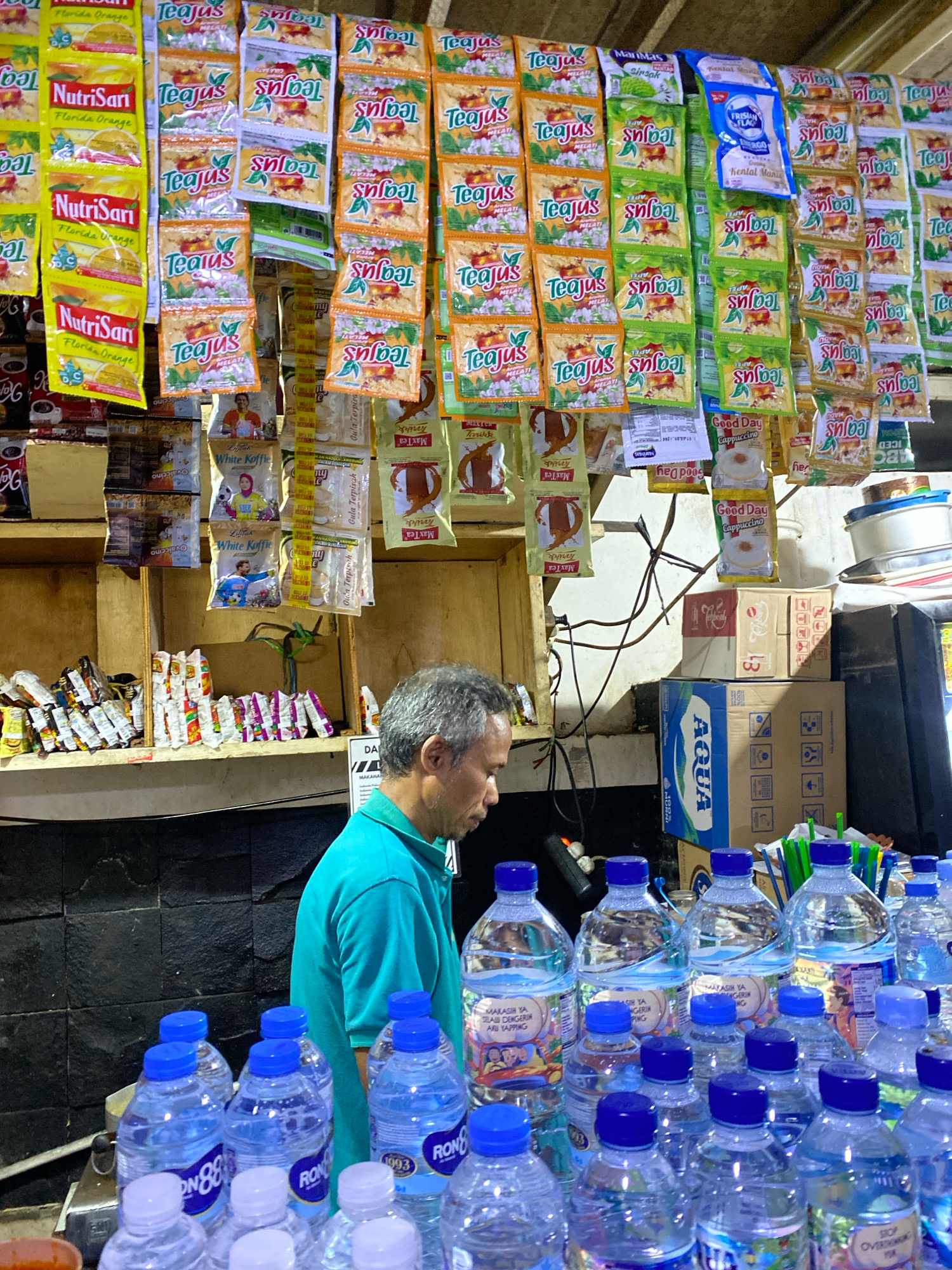 A middle-aged man with graying hair, wearing a teal polo shirt, stands behind a counter in a dimly lit store. The counter is filled with numerous plastic bottles of water, most with blue caps and clear liquid, some displaying "RON 88" branding. Behind the man, shelves are laden with a variety of bagged food and drink products, including brightly colored packets of NutriSari and Teajus, as well as boxes of "Aqua" bottled water. The background reveals wooden shelves and dark tiled walls, suggesting a small convenience store or warung. The overall impression is one of a typical Indonesian roadside stall or small shop.