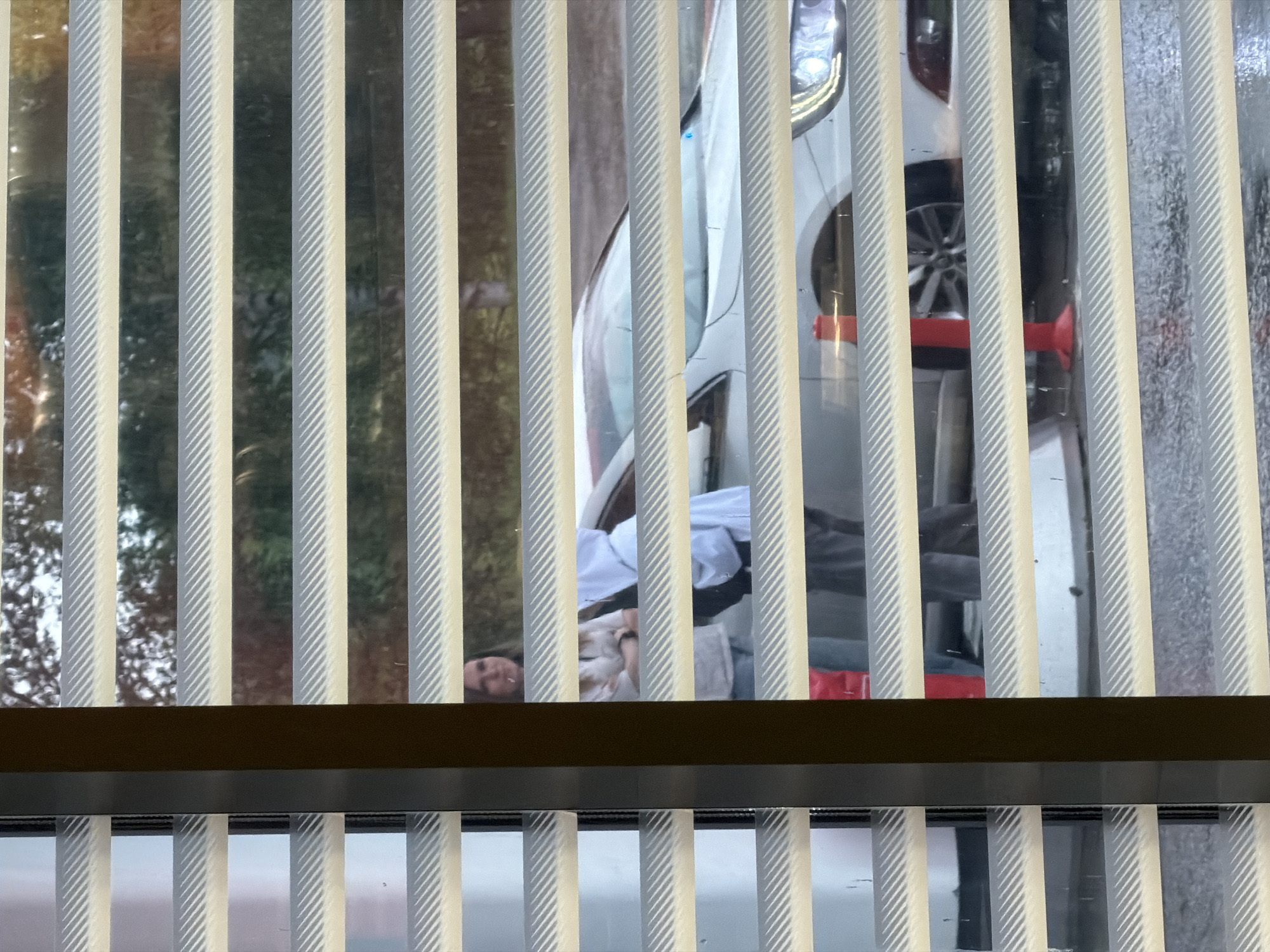 This is a vertical-format, medium-angle, eye-level shot taken through vertical blinds or louvers. The blinds are pale yellow and cream-colored with a textured, ribbed surface. They create a pattern of vertical stripes across the entire image, partially obscuring the scene behind them.
The scene behind the blinds appears to be an outdoor setting, likely a street or a parking area, with trees and foliage visible to the left. A white car is the most prominent object in the background. The car's headlights, grill, and part of its wheel are visible. There are also some red and orange objects on the ground near the car, possibly traffic cones or safety equipment.
A person is visible in the middle ground, behind the blinds. They are lying down, with their head and upper body visible. They are wearing a white garment that appears to be a shirt or a robe. Their face is partially visible, and they seem to be looking upwards. The lighting suggests it might be daytime, possibly late afternoon or early evening, given the soft, diffused light.
The overall impression is one of observation or a clandestine view. The blinds act as a barrier, giving a sense of privacy or separation between the viewer and the scene. There is no visible text in the image. The location context is Krilovo, Russia.