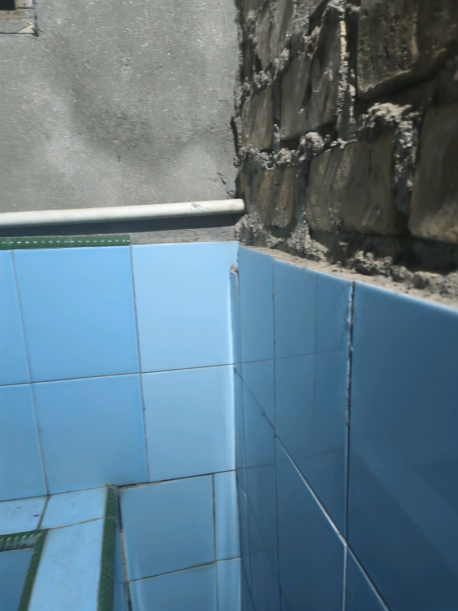 The image shows an interior corner where two walls meet. One wall is covered in light blue square tiles and the other is constructed of rough, irregular stones cemented together. A horizontal white pipe runs along the base of the stone wall, above the tiled wall. The tiles are also present along the base of the stone wall, suggesting a boundary between the two wall types. The stone wall has visible mortar and rough edges. The tiled wall appears to be a standard bathroom or shower enclosure. The lighting suggests an indoor environment, possibly with a window or overhead light source. The scene is devoid of any individuals. The location is unknown, Indonesia.