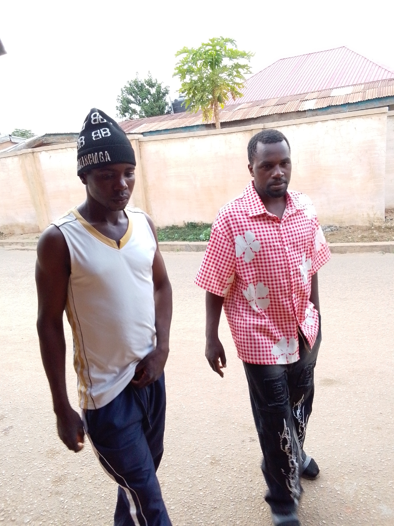 The image captures two men standing outdoors, likely on a street or unpaved path in Jos, Nigeria. The ground is a light, dusty color. In the background, there is a low, light-colored wall or fence, behind which are visible corrugated metal roofs (one reddish-brown, one grey) and several green trees under a bright, possibly overcast sky.
The man on the left wears a black knitted beanie featuring the letters "BB" repeated and the brand name "BALENCIAGA" in white text. He is dressed in a white sleeveless tank top with a gold-trimmed V-neck and gold stripes along the sides. He wears dark blue athletic pants with white side stripes. His expression is neutral as he looks slightly downwards.
The man on the right wears a short-sleeved, red and white checkered shirt adorned with large white floral or leaf-like patterns. He is wearing dark, possibly distressed jeans with white graphic designs on the legs and dark shoes or sandals. He also maintains a neutral expression, looking generally forward.
Both men have dark skin, short dark hair, and some facial hair. They appear to be casually standing or walking, with their hands either in their pockets or resting at their sides. The overall setting suggests a daytime scene in a residential or semi-urban environment.