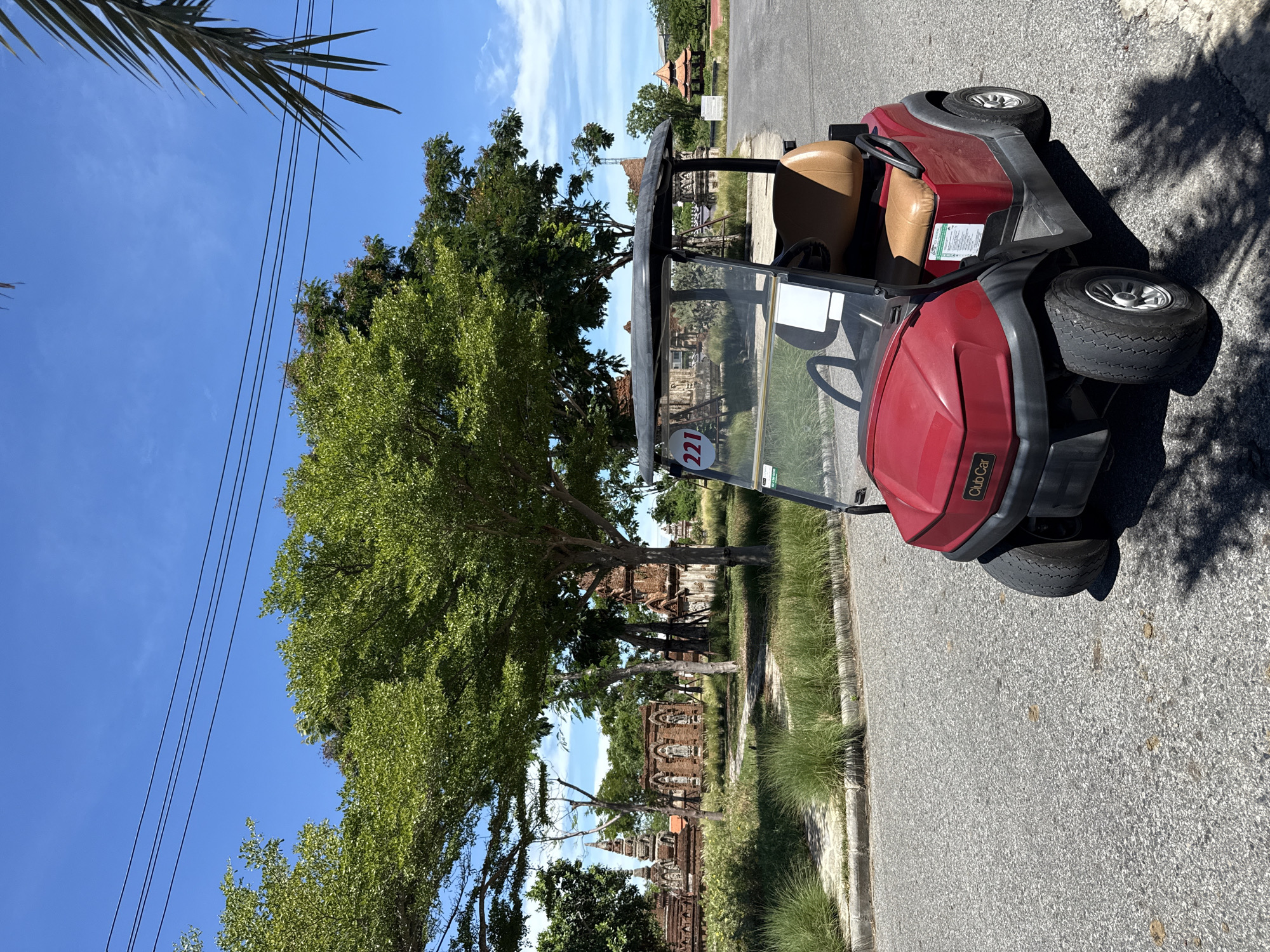 The image shows a red Club Car golf cart parked on an asphalt road in Bang Pu Subdistrict Municipality, Thailand. The cart is angled slightly towards the right side of the frame, with its front facing the camera. The number "221" is visible on the side of the cart. Behind the cart, there are trees and the ruins of what appears to be ancient temples or structures. The sky is blue with some clouds. Power lines are visible in the upper left corner of the image. The lighting suggests it is daytime, with shadows cast by the cart and trees. The environment appears to be a tourist or historical site.