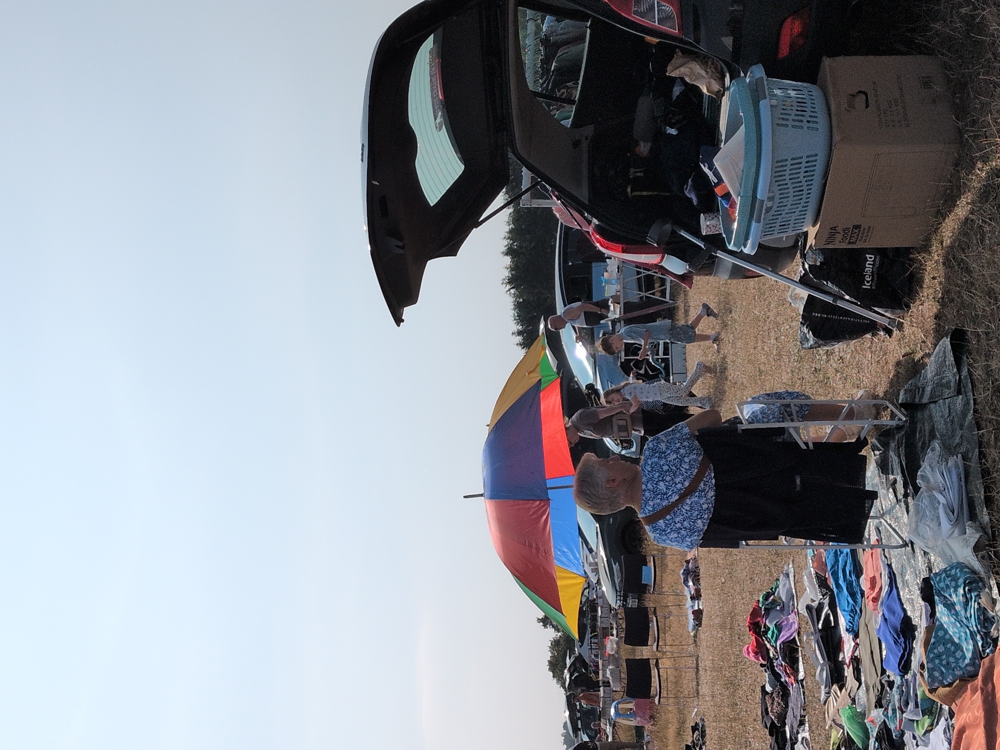 The image depicts an outdoor car boot sale on a sunny day in Claverley, United Kingdom. In the foreground, a woman sits in a chair under a large, multi-colored umbrella, facing away from the camera. She is wearing a blue and white patterned top and a dark cardigan. In front of her, on the ground, are piles of folded clothes and blankets laid out on tarpaulins, suggesting a stall selling second-hand items.
To the right of the woman, a dark-colored car with its boot open is parked. Inside the boot, various items are visible, including clothing and bags. A blue laundry basket sits on top of a cardboard box next to the car. A long, silver pole is propped against the car.
In the middle ground, several cars are parked, and other people are browsing or setting up stalls. A young boy in a light blue t-shirt and shorts is walking past the cars, looking down at something in his hands. A young girl in a white top and patterned leggings is walking towards a man who is wearing a dark shirt and holding a brown bag. Two black folding chairs are set up near another car. A small, red and blue toy vehicle is also visible.
In the background, more cars and people can be seen, as well as trees lining the horizon under a clear, pale blue sky. The ground is covered in dry grass and patches of earth, indicating a field or open area. The overall scene suggests a busy and active community event.