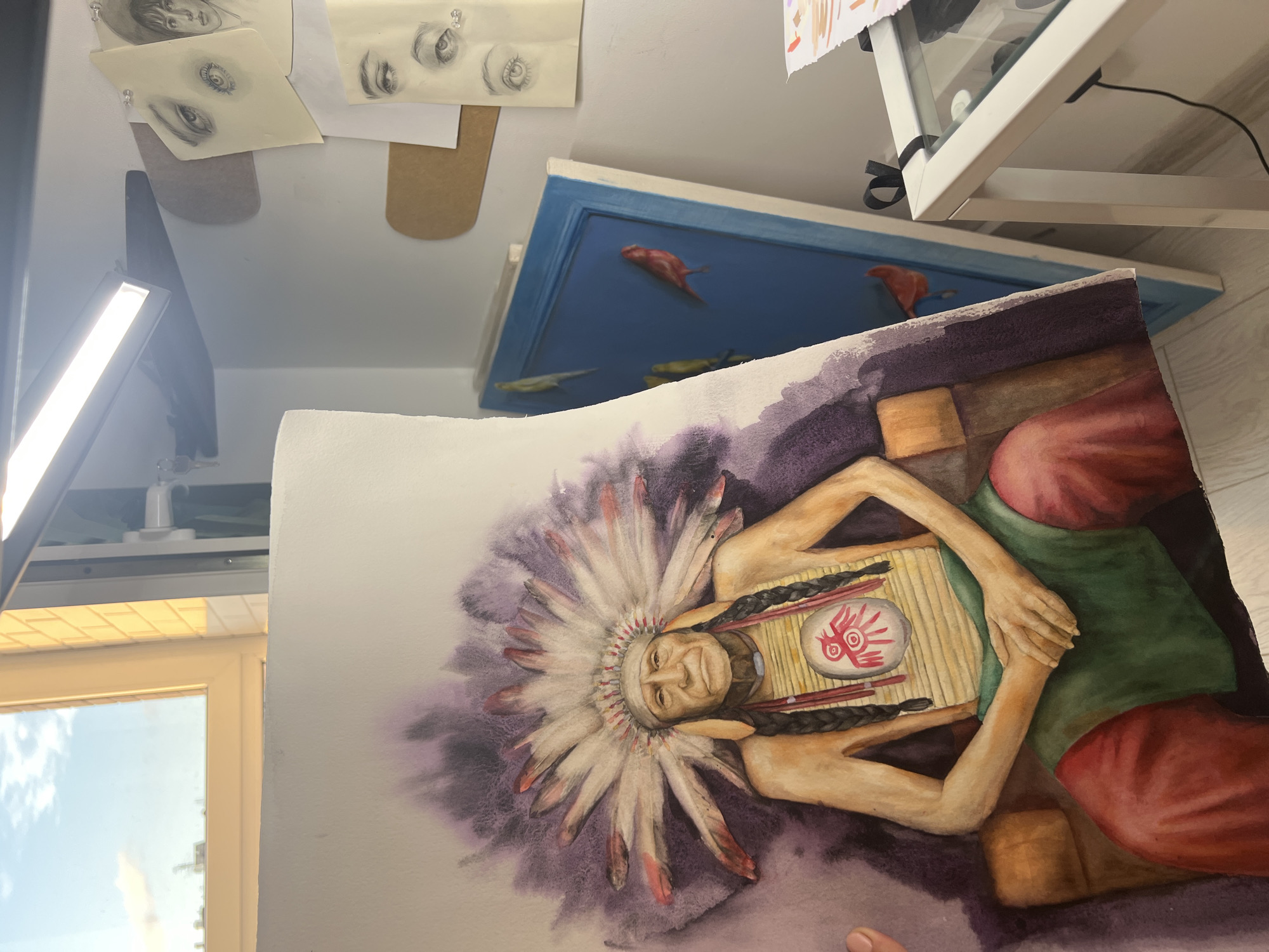 The image shows a watercolor painting of a Native American chief wearing a headdress and traditional attire. The painting is held in the foreground, partially obscuring a blue canvas with painted birds and a white table with glass shelves in the background. There are also several drawings of eyes pinned to the wall. The scene appears to be an artist's studio or workspace, with natural light coming from a window and artificial light from a lamp. The location is not specified beyond "Unknown, Unknown."