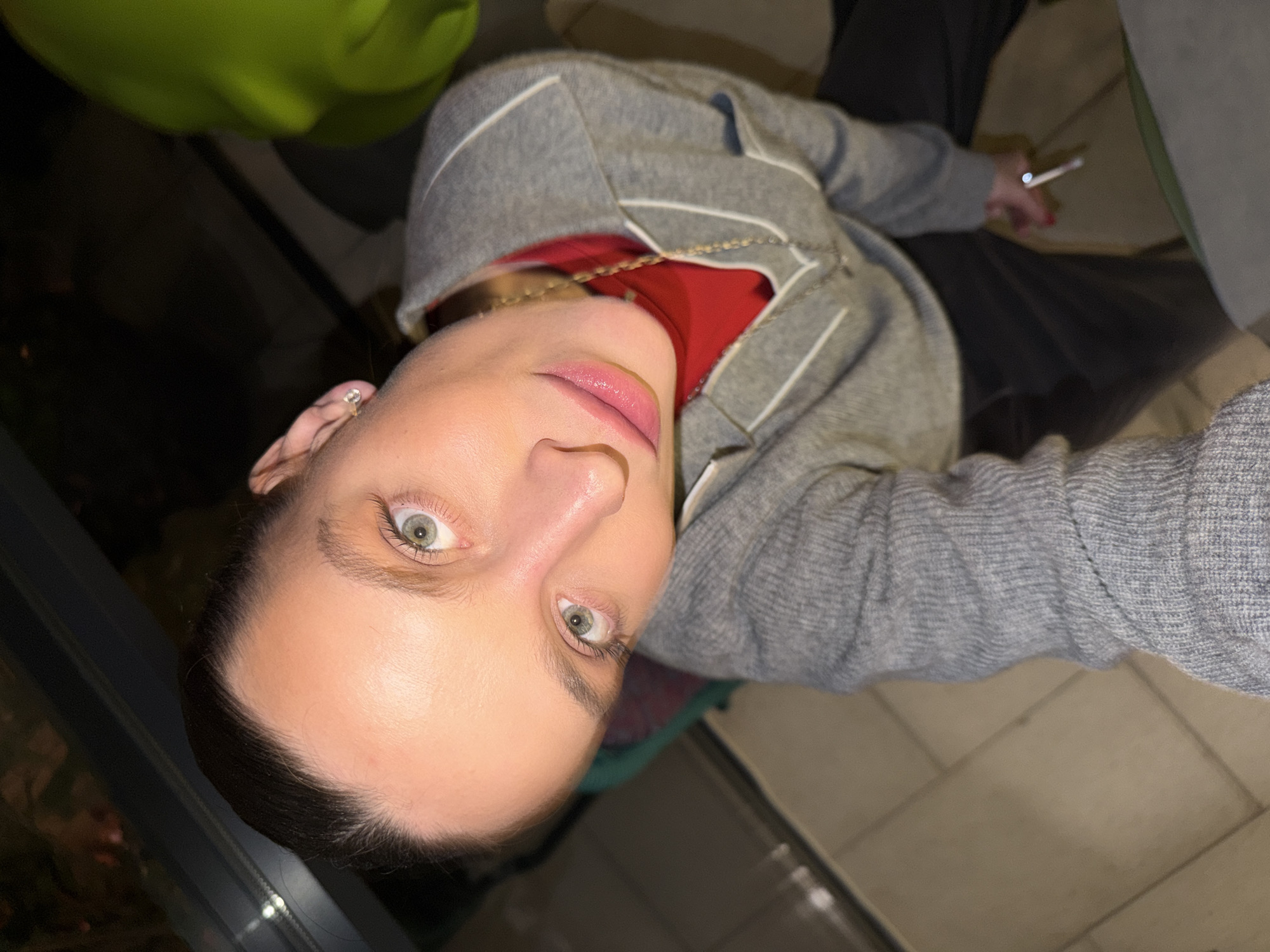 A person with close-cropped dark hair and light eyes, wearing a gray cardigan over a red top and gold chain necklace, is holding a cigarette in their right hand. They are sitting in a chair with a green cushion in what appears to be an indoor setting. The background shows a tile floor and a glimpse of what could be a window or glass door. There is no visible evidence to confirm a specific location in Budva, Montenegro.