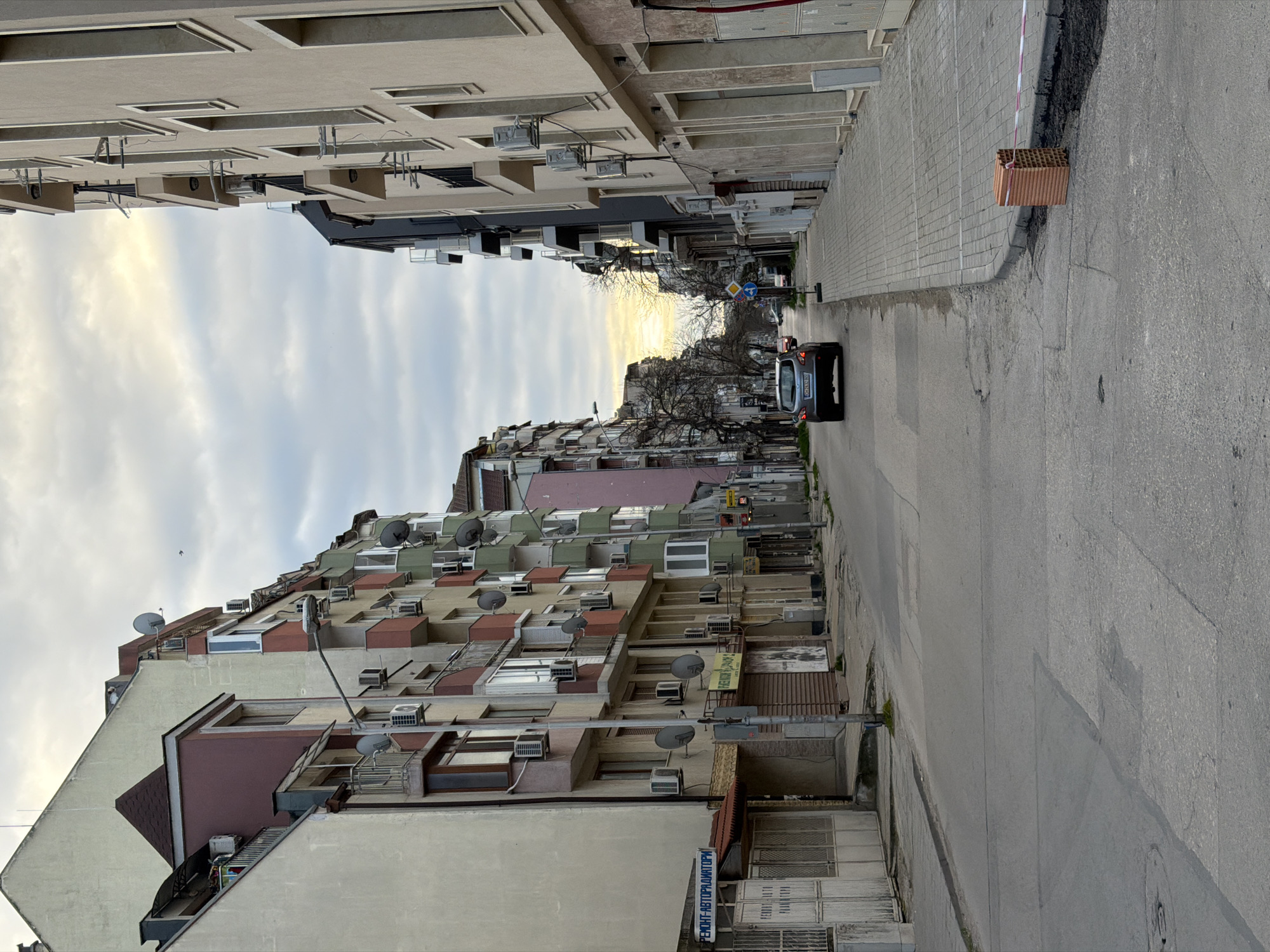 A street scene in Varna, Bulgaria, shows a narrow street lined with buildings on either side. The buildings are multi-storied and appear to be residential, with visible air conditioning units and satellite dishes. The street is paved and leads to a distant intersection where traffic signs are visible. A dark-colored car is driving down the street. A stack of bricks is on the right side of the street with some red and white tape next to it. The sky is overcast with light breaking through in the distance. A sign with Cyrillic text is visible on one of the buildings. The scene is shot from a ground-level perspective.