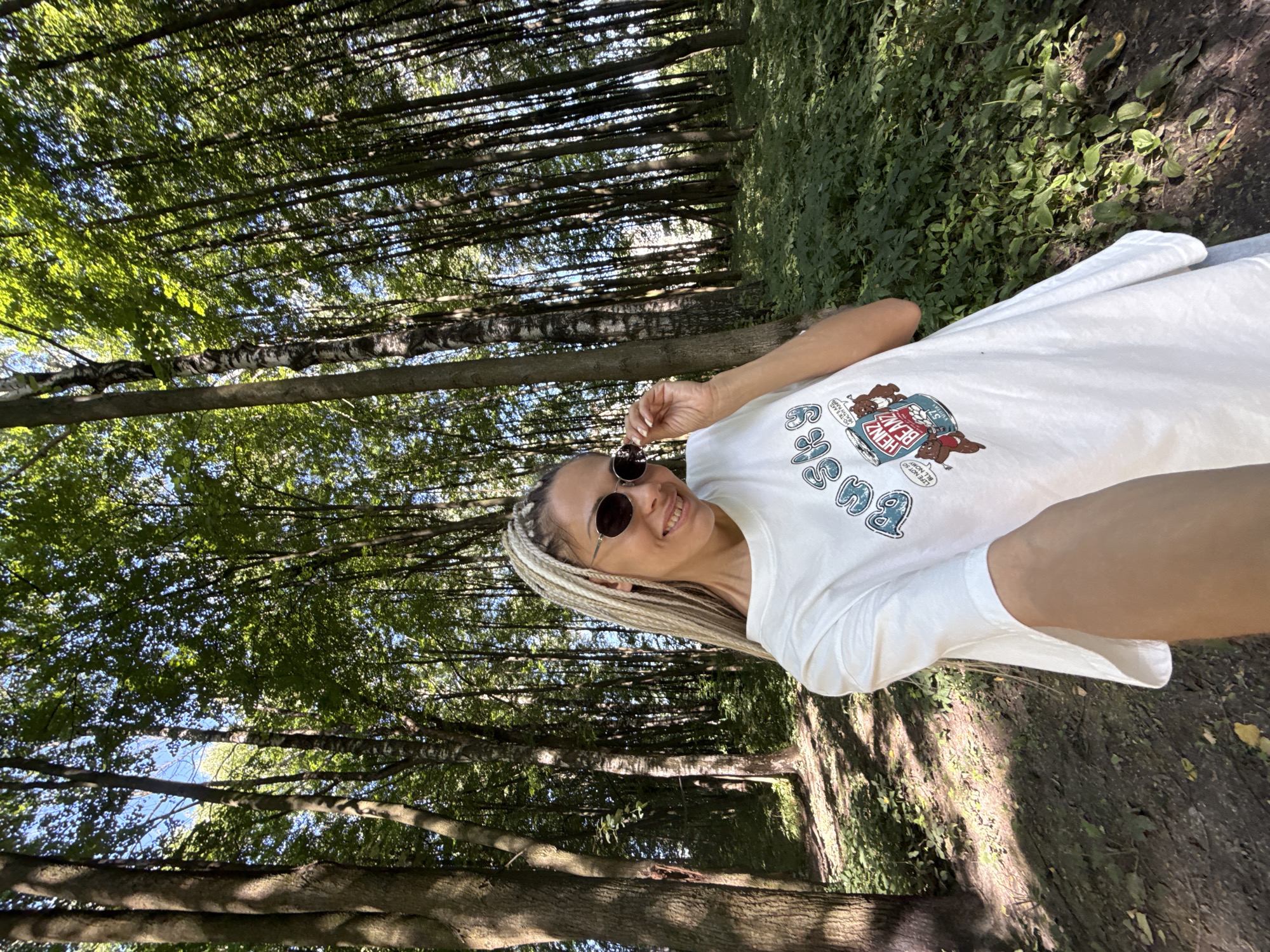A medium shot captures a woman with blonde braided hair and sunglasses taking a selfie in a forest. She is smiling and adjusting her sunglasses with her right hand. She is wearing a white t-shirt with a graphic print of a Heinz baked beans can and cartoon bears. The background is a dense forest with tall trees, lush green undergrowth, and dappled sunlight filtering through the leaves. The ground is covered in dirt and fallen leaves. The location is identified as Rusanovo, Russia.