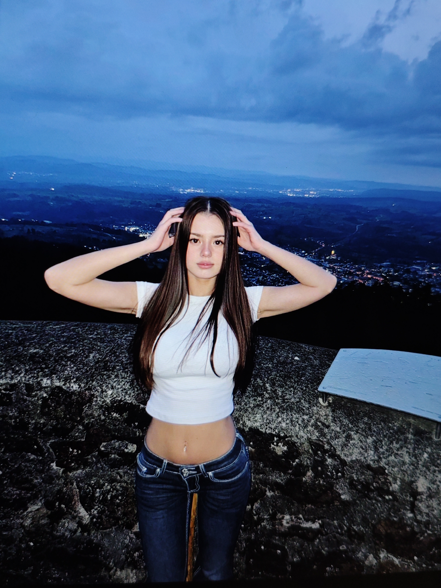A young woman stands on a rocky outcrop overlooking a city at twilight. She has long, dark brown hair and is wearing a white crop top and blue jeans. Her hands are raised to her head. The city lights twinkle in the distance below, and the sky is a deep blue with hints of clouds. The environment appears to be a natural landscape with a view of urban sprawl. The specific location is not identifiable as Baku City, Azerbaijan, based on visual cues alone.