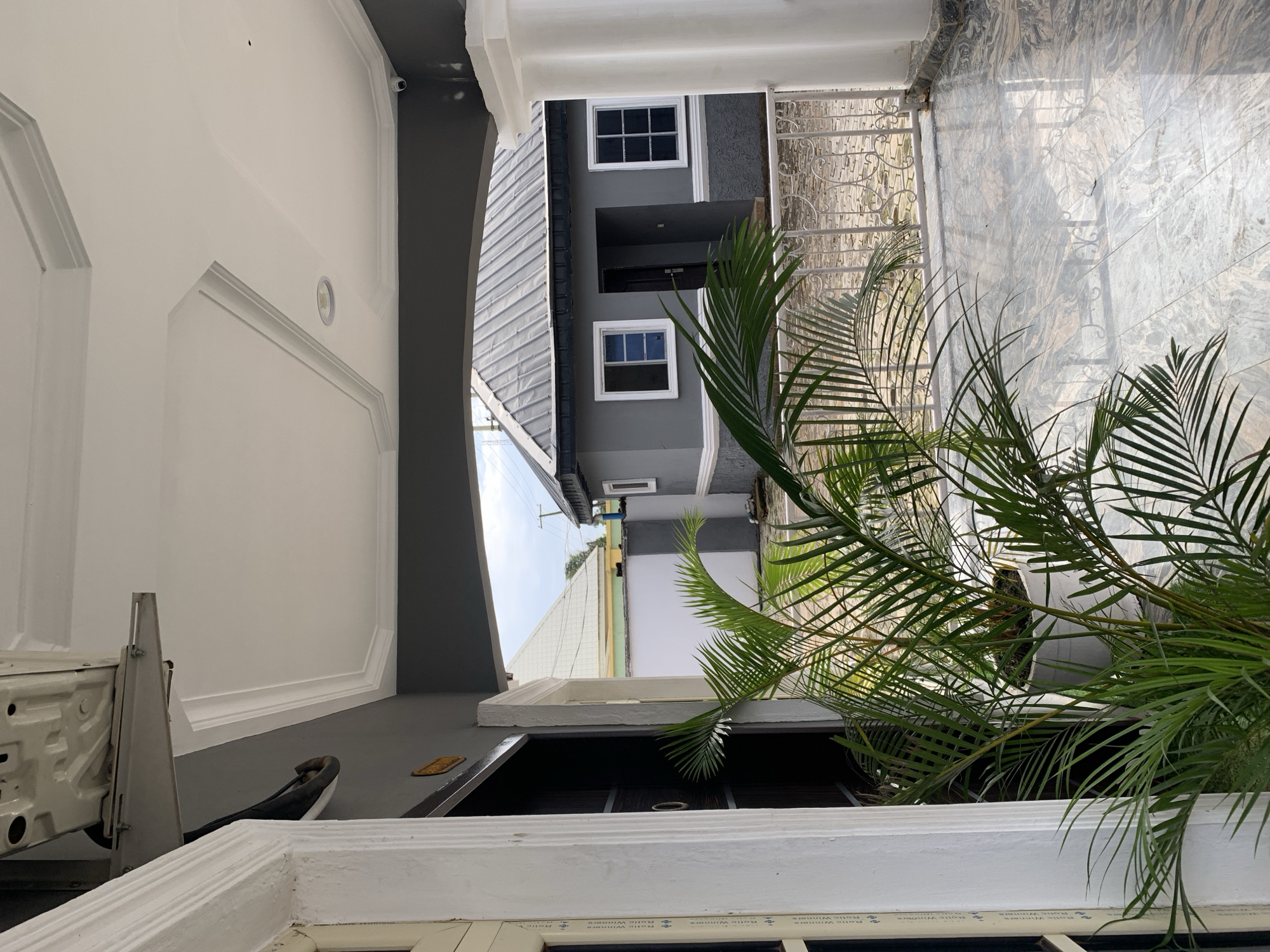 A wide-angle exterior shot captures a covered porch with a potted palm plant in the foreground. The porch features a marbled floor and a white column on the right. Beyond the porch, a gray building with a silver metal roof is visible, featuring two white-framed windows and an open doorway. A white decorative metal fence separates the porch area from the building. In the background, other buildings and utility poles suggest a residential or commercial area within the city of Gura Top, Nigeria. The sky is overcast.