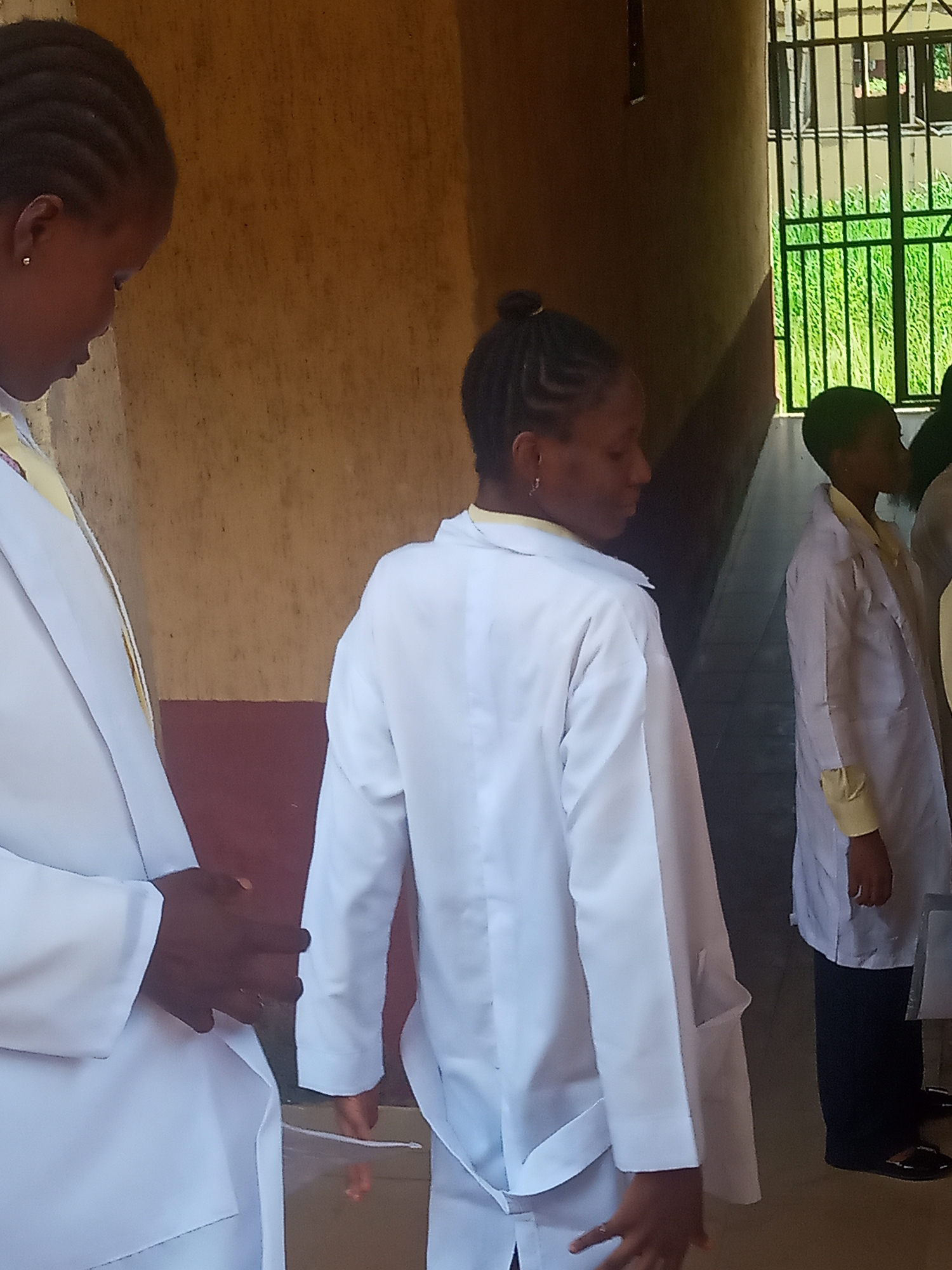This image depicts a group of young women or girls, likely students or trainees, standing in what appears to be a corridor or covered walkway in Nkwelle, Nigeria. All subjects are wearing white lab coats over light yellow or cream-colored shirts.
The individual in the foreground on the left is seen from the chest up, with braided hair, facing slightly away from the camera with her head bowed. Her hands are clasped in front of her. In the middle of the frame, another individual with braided hair tied in a bun stands facing to the right, also looking downwards. Further back on the right, a third person in a lab coat is visible, facing in the same direction.
The setting features a plain, light brown wall behind the subjects, with a darker reddish-brown section along the bottom. Bright natural light from the right side of the frame suggests it is daytime. A window with dark metal bars is visible on the far right, offering a view of vibrant green foliage outside. The overall scene suggests a formal or educational environment, with the individuals standing attentively. No visible text is present.