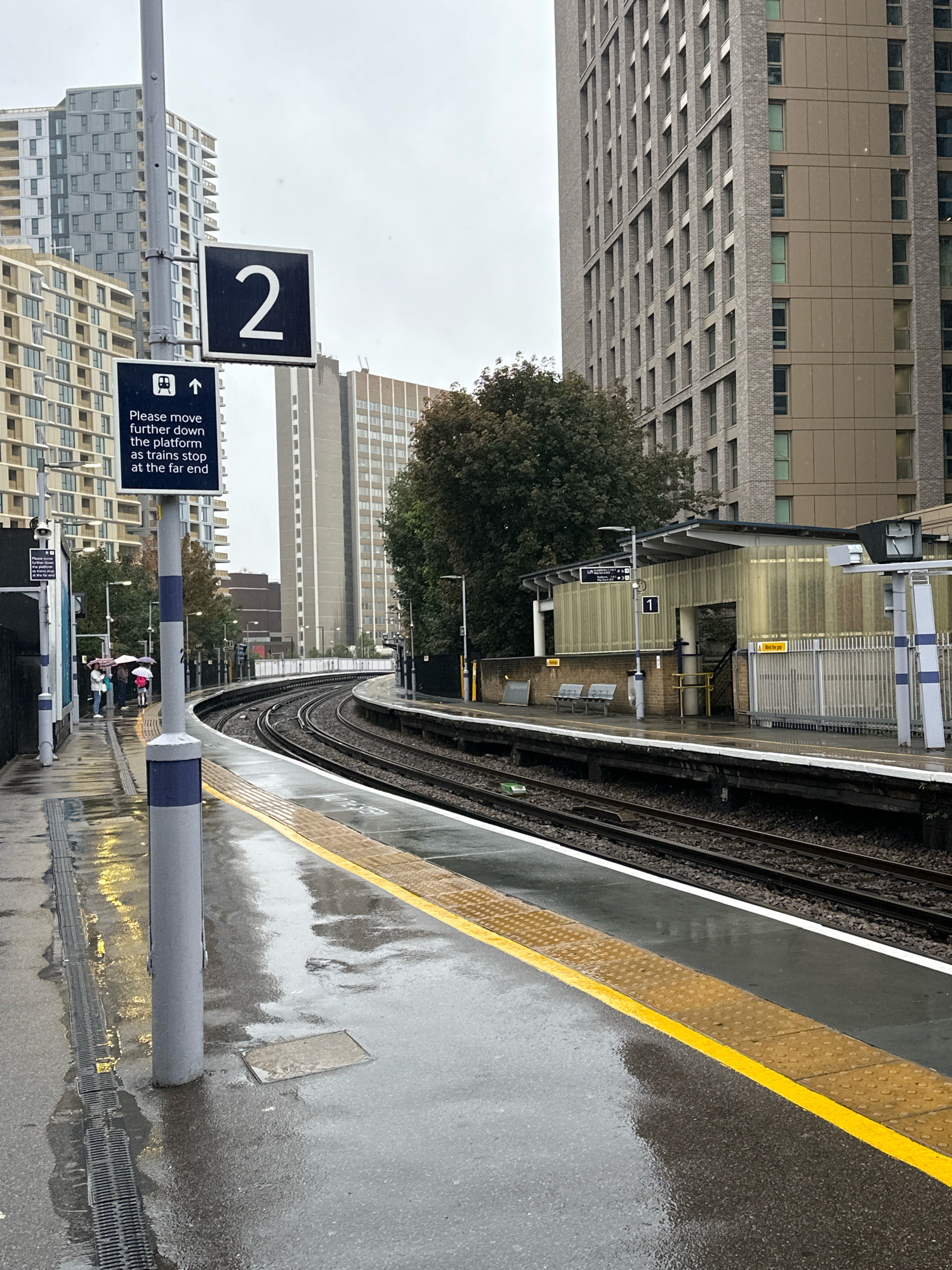 The image depicts a rain-slicked platform at a train station, likely in London, United Kingdom. The platform is wet and reflective, with puddles visible on the ground. A yellow tactile paving line runs along the edge of the platform, indicating a safety warning for visually impaired passengers.
In the foreground, a grey pole with blue and white banding stands prominently. A sign attached to this pole displays a large white number "2" within a black square, indicating platform 2. Below this, another sign, also with a blue border, features an icon of a train and an upward-pointing arrow, accompanied by text that reads "Please move further down the platform as trains stop at the far end."
Several people are visible on the platform in the middle ground, holding umbrellas and appearing to wait for a train. They are situated further down the platform, consistent with the advisory sign. The train tracks curve away from the viewer into the distance.
In the background, modern multi-story buildings with a grid-like facade rise above the station structures. A tree with dense foliage is also visible between the station building and the taller structures. The sky appears overcast, contributing to the gloomy atmosphere and the wet conditions. The station itself has a covered waiting area with benches, a metal fence, and translucent yellow panels. A sign with the number "1" is visible on the station building, indicating platform 1.