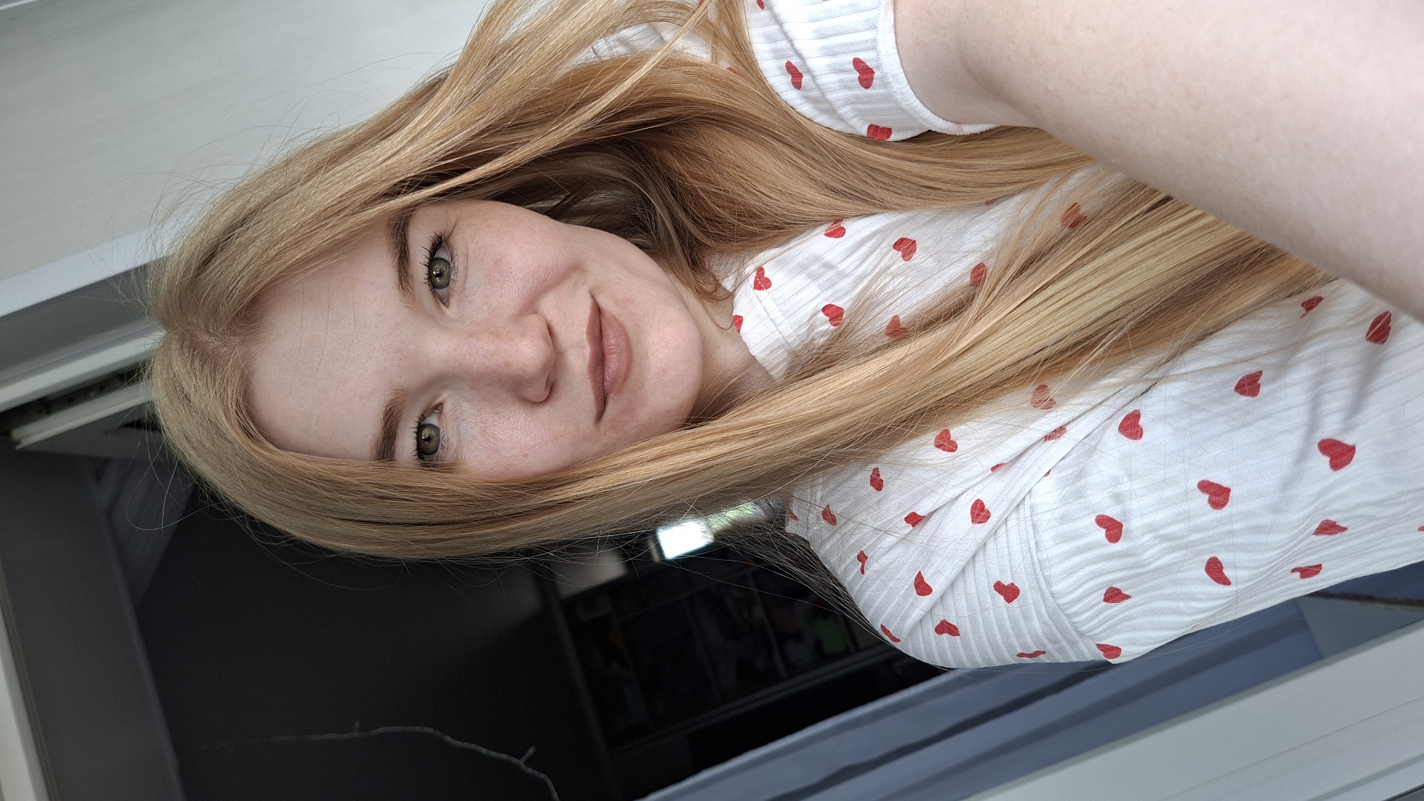 A selfie of a fair-skinned woman with long blonde hair wearing a white shirt with red heart patterns. She has green eyes and freckles. The background shows a window with a dark interior beyond. No information indicates this image was taken in Chaikovsky, Russia.