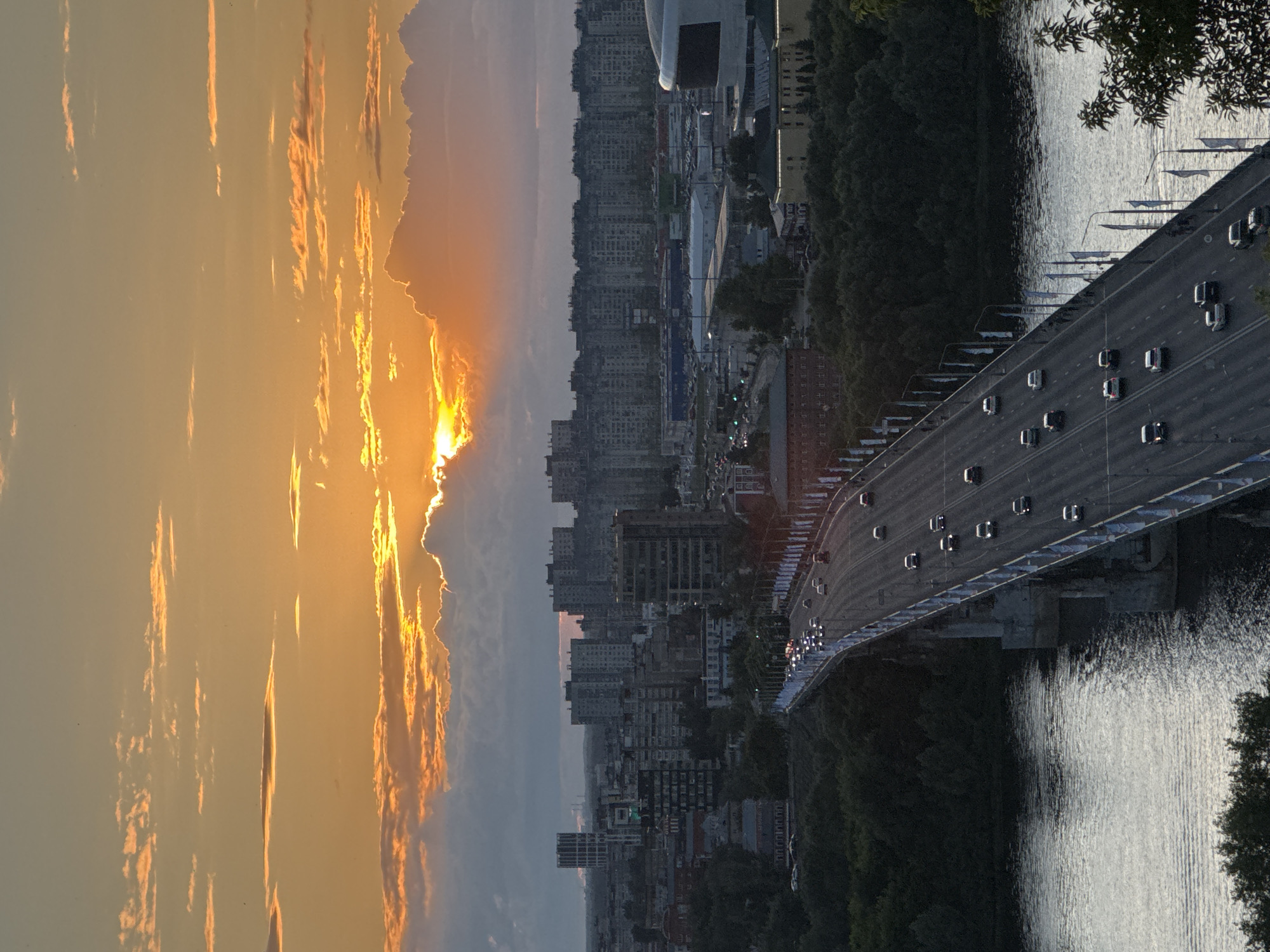 The image captures a sunset over the city of Nizhny Novgorod, Russia. A wide bridge, spanning a river, dominates the foreground, with traffic visible on its lanes. The bridge is adorned with flags and banners. On either side of the river, lush green trees line the banks. Beyond the trees, the city skyline unfolds, characterized by numerous apartment buildings and industrial structures. The setting sun, partially obscured by clouds, casts a warm, orange glow across the sky, illuminating the scene with dramatic light. The overall impression is one of a serene urban landscape at dusk.