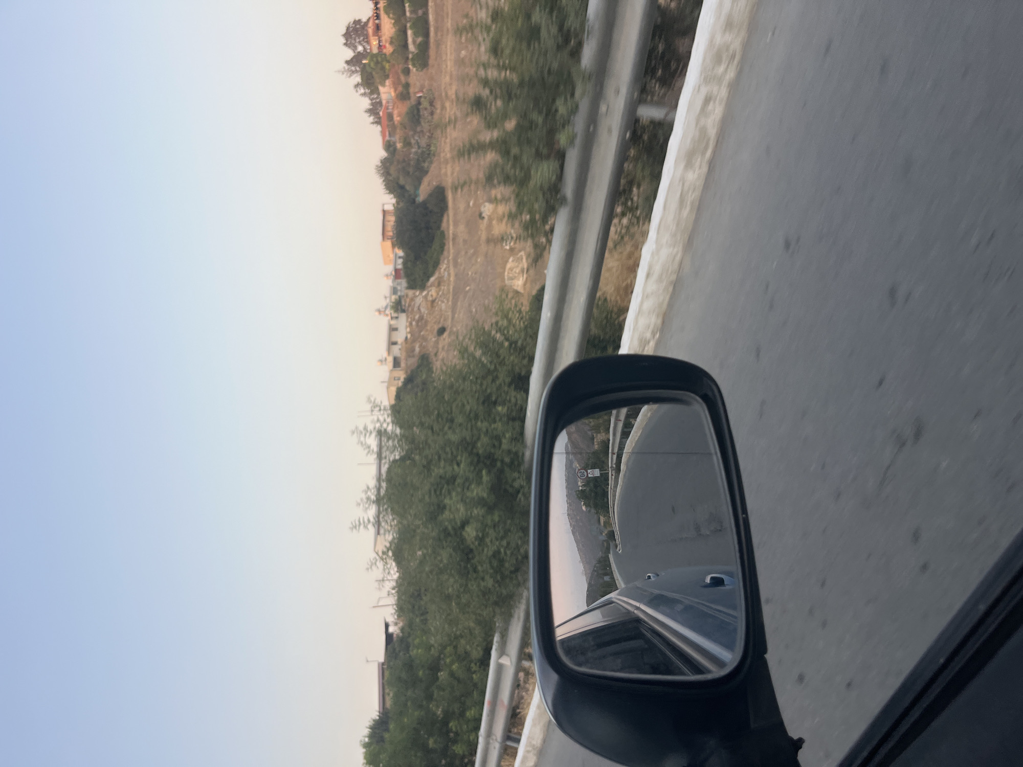 The image depicts a view from a moving vehicle, capturing a roadside scene in the Armenochori Community, Cyprus. The foreground is dominated by the side mirror of the car, reflecting a winding road that curves into the distance. The mirror also shows the side of the vehicle and a glimpse of the sky.
Outside the mirror, to the right of the frame, a road with a white concrete barrier runs alongside. Beyond the barrier, the landscape is characterized by dry, brown earth dotted with sparse vegetation and trees. In the background, several buildings, some appearing residential with terracotta roofs, are nestled on a hillside. The sky above is clear and light blue, transitioning to a softer hue near the horizon, suggesting it might be late afternoon or early morning. The overall impression is a tranquil, rural setting on a clear day.