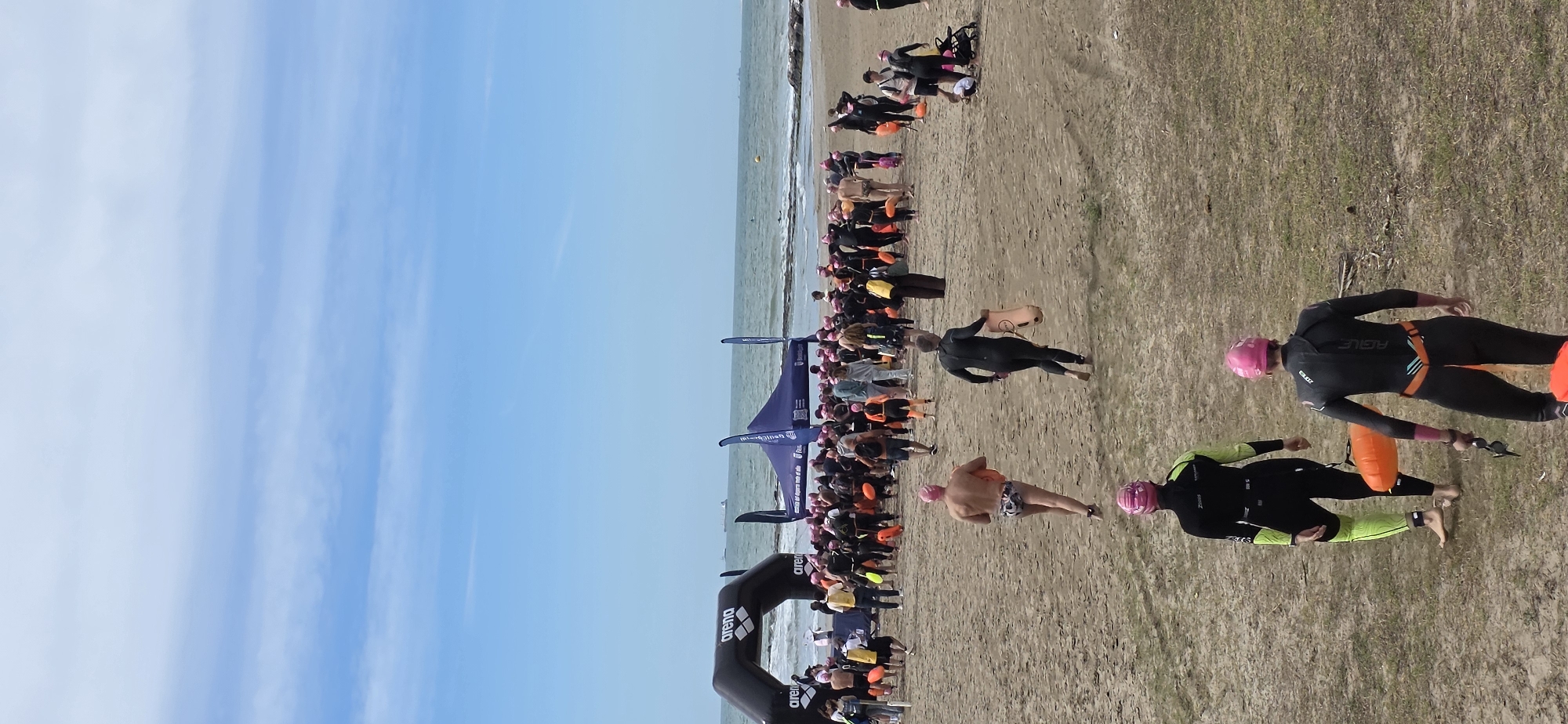 The image depicts a triathlon event on the beach in Benicàssim, Spain. Swimmers are exiting the water and running onto the sand, some wearing wetsuits, pink swimming caps, and carrying orange buoys. An "Arena" branded inflatable archway marks the transition area. Spectators and event staff are visible along the beach. The sky is blue with scattered clouds, and the sea is calm.
