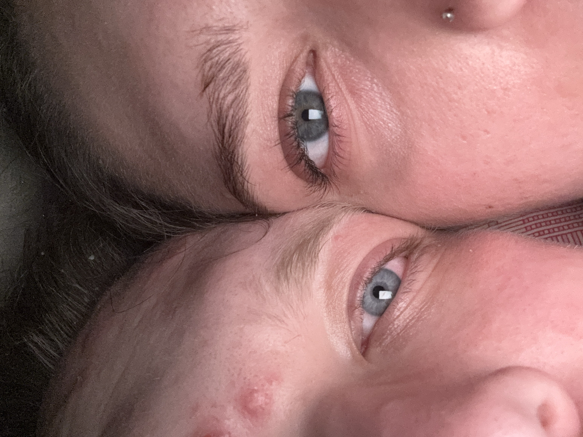 The image is a close-up, low-angle, indoor photograph of two individuals' faces, positioned so that their eyes are nearly touching. The lighting is warm and appears to be from an artificial source.
On the left side of the frame, a portion of a face is visible, focusing on the left eye. This individual has pale skin with some redness and a visible pimple or blemish on the forehead. The left eye is open, revealing a blue iris. The eyebrows are light brown and somewhat sparse.
On the right side of the frame, the right eye of another individual is prominently displayed. This person also has pale skin, with some light redness. The right eye is open, showing a blue-gray iris with a distinct reflection of a light source in the pupil. The eyebrows are darker and fuller than the other individual's. A small nose piercing is visible on the cheekbone.
Both individuals appear to be close together, suggesting an intimate or shared moment. The background is dark and out of focus, with a hint of texture that could be fabric, possibly a pillow or bedding. There are no discernible elements that would indicate a specific location within the city of Saratov, Russia. The scene is forensic in nature, offering a detailed view of facial features and skin texture.