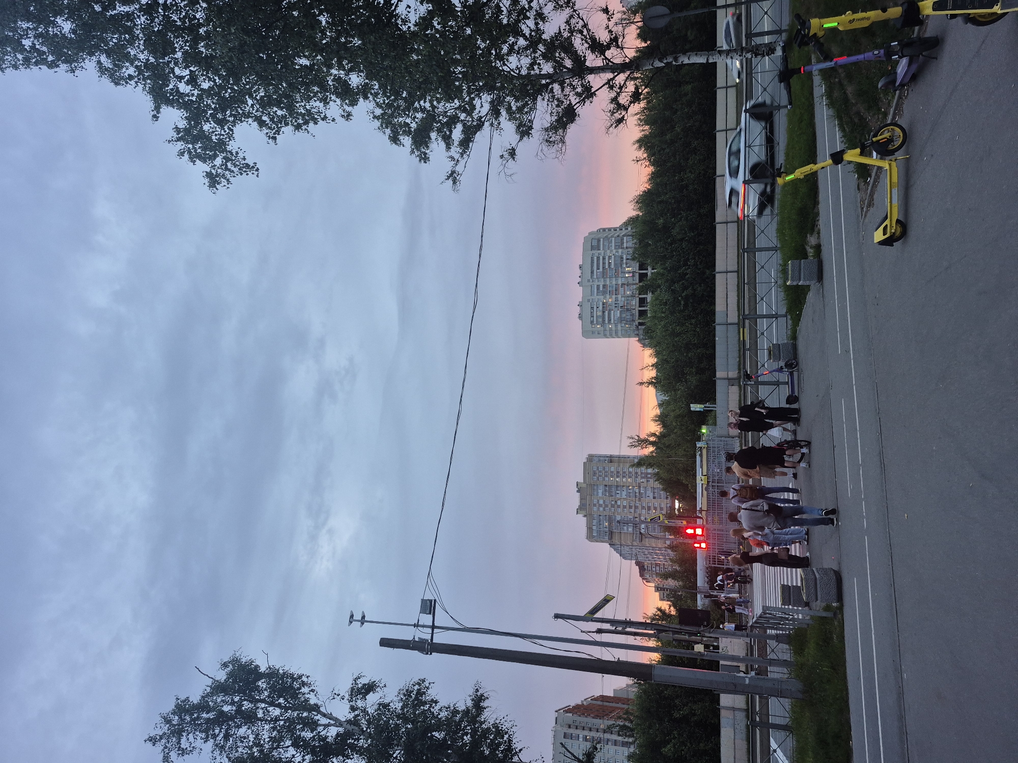 The image depicts a street scene in Saint Petersburg, Russia, during twilight. The sky is filled with a gradient of blue and pink hues, indicating sunset or sunrise. On the right side of the image, several parked electric scooters are visible, with yellow and purple models prominent. A white car is in motion on the road in the background, blurred due to its speed. On the left side of the image, people are gathered near a crosswalk, waiting to cross the street. Some individuals are standing, while others appear to be walking. Traffic lights are red, indicating that vehicles should stop. Tall apartment buildings and lush green trees form the backdrop of the scene. A street lamp and electrical wires are also visible, characteristic of an urban environment. The overall atmosphere is that of a quiet evening in a residential area of Saint Petersburg.