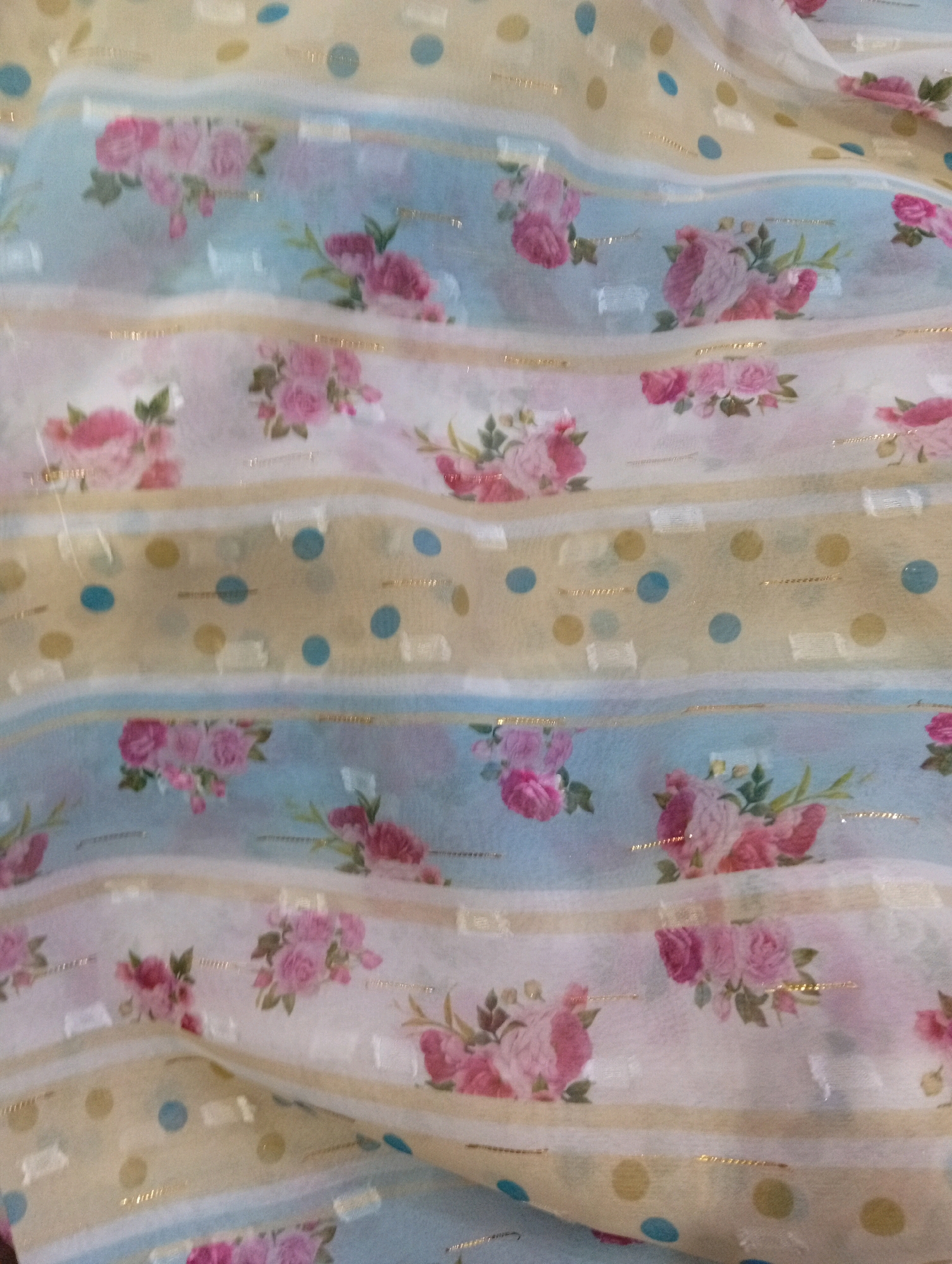 The image shows a close-up, overhead view of a fabric with a floral and polka dot pattern. The fabric is draped, creating folds and shadows. The pattern features horizontal stripes of varying widths. Some stripes are light blue with scattered pink and red roses. Other stripes are off-white with gold metallic threads and scattered small white square patterns. There are also wider yellow stripes with a pattern of gold and blue polka dots. The overall impression is of a light, airy fabric, possibly a dress or scarf. The image does not contain any elements that would allow for the identification of its location in Larkana, Pakistan.