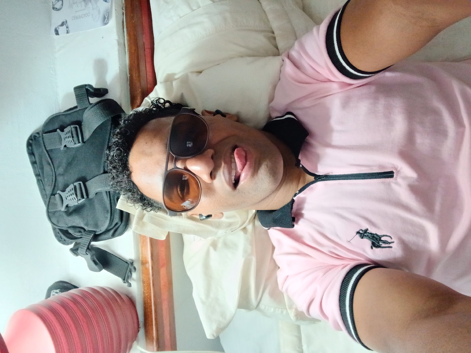A man is lying on a bed, wearing sunglasses and a pink polo shirt with black and white trim. His tongue is sticking out. Next to the bed is a black backpack and a red cylindrical object. The location is likely Porlamar, Venezuela. There is also a white wall with what looks like a document/poster on it.