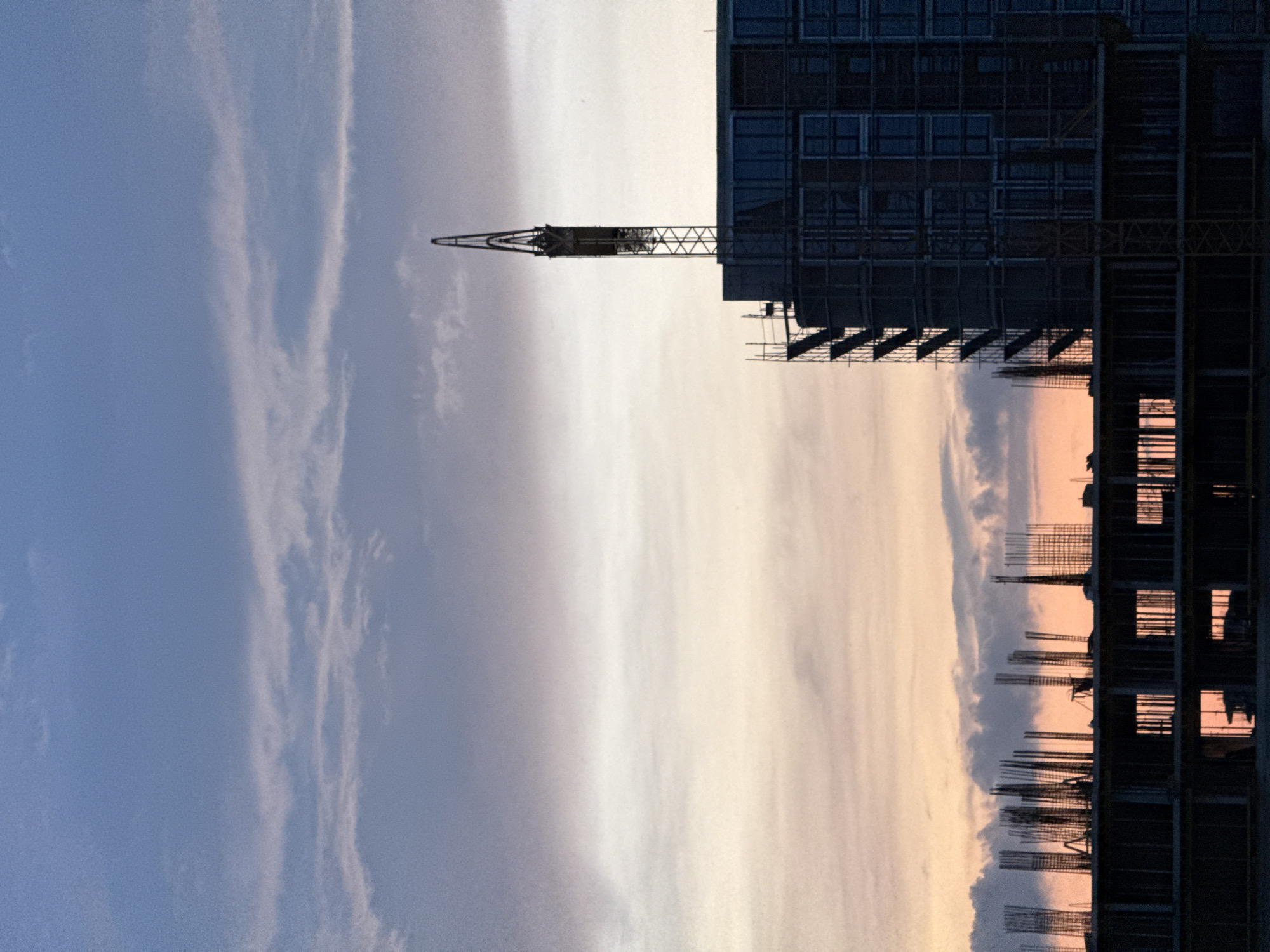 A construction site is depicted under a dramatic, colorful sky, likely at sunrise or sunset. The image shows a large building under construction, silhouetted against the bright backdrop. To the left of the main building, a tall tower crane is also visible in silhouette, its lattice structure clearly outlined.
The building exhibits various stages of construction, with visible rebar cages extending upwards, suggesting ongoing concrete work. Sections of scaffolding can be seen clinging to the dark facade of the partly completed structure, which also features dark, window-like openings. The overall impression of the building is one of structural complexity and robust engineering in progress.
The sky is a prominent feature, displaying a striking gradient of colors. On the left, it transitions from a soft blue to a lighter, almost white hue towards the center, then to warm orange and pink tones on the right, indicative of either dawn or dusk. Wispy, elongated clouds stretch across the sky, catching the light and adding texture. The lighting is strong backlighting, causing the construction elements to appear as dark, sharply defined silhouettes against the luminous sky.
No people are visible in the frame, and there is no visible text. The scene captures a moment of quiet activity or preparation at a construction site, possibly in Burgas, Bulgaria, given the provided location context. The atmosphere is one of serene industrial progress set against a beautiful natural backdrop.