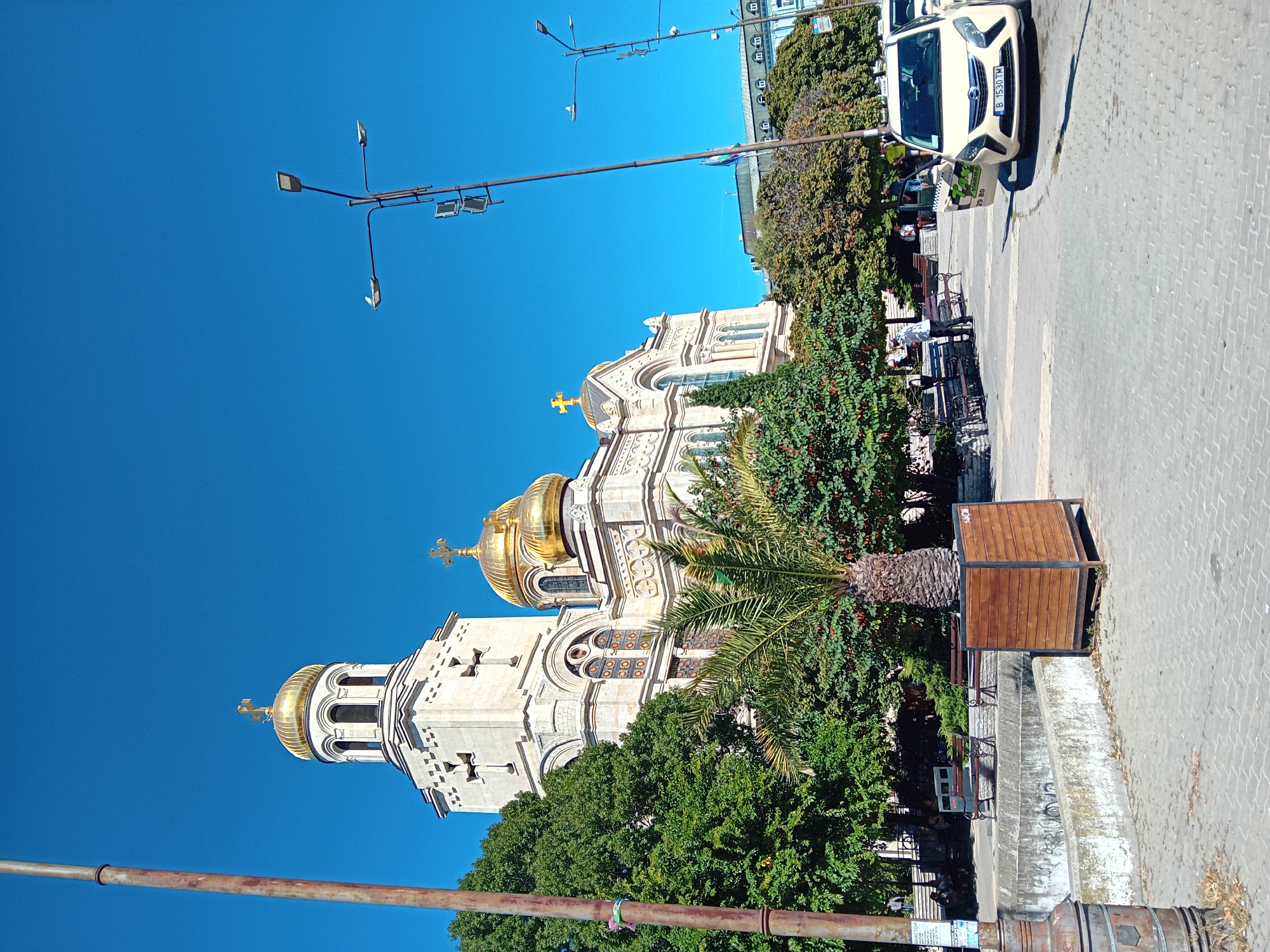 The image captures the Cathedral of the Assumption of the Theotokos in Varna, Bulgaria, on a bright, clear day. The cathedral, a prominent landmark with its golden domes and white stone facade, is partially obscured by lush green trees. In the foreground, a paved plaza features park benches, a large wooden planter with a palm tree, and a light-colored taxi with its doors open. Streetlights with solar panels are visible, along with a weathered metal pole in the left foreground. Several individuals are scattered throughout the scene, some sitting on benches and others standing, indicating a public space with moderate activity. The sky is a vibrant, cloudless blue, and the overall impression is one of a sunny day in a well-maintained urban environment.