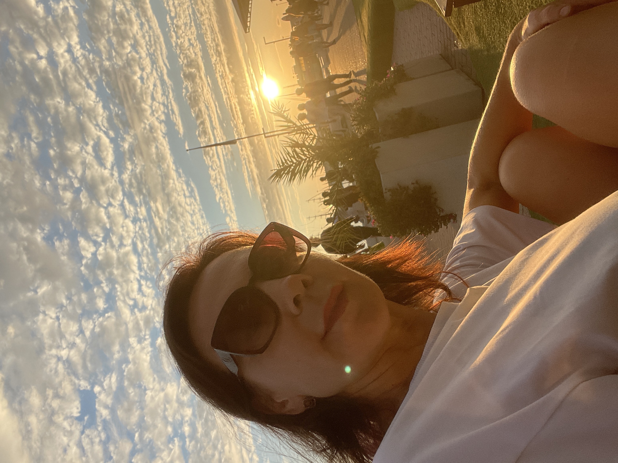 A woman wearing sunglasses and a white t-shirt is taking a selfie outdoors during sunset. The sun is visible in the background, casting a warm glow on the scene. In the background, there are people walking along a promenade with palm trees and potted plants. The sky is filled with scattered clouds, illuminated by the setting sun. The woman is sitting down, with her legs crossed. The location appears to be a waterfront promenade, possibly in Sochi, Russia, given the presence of palm trees and the general ambiance of a seaside resort town.