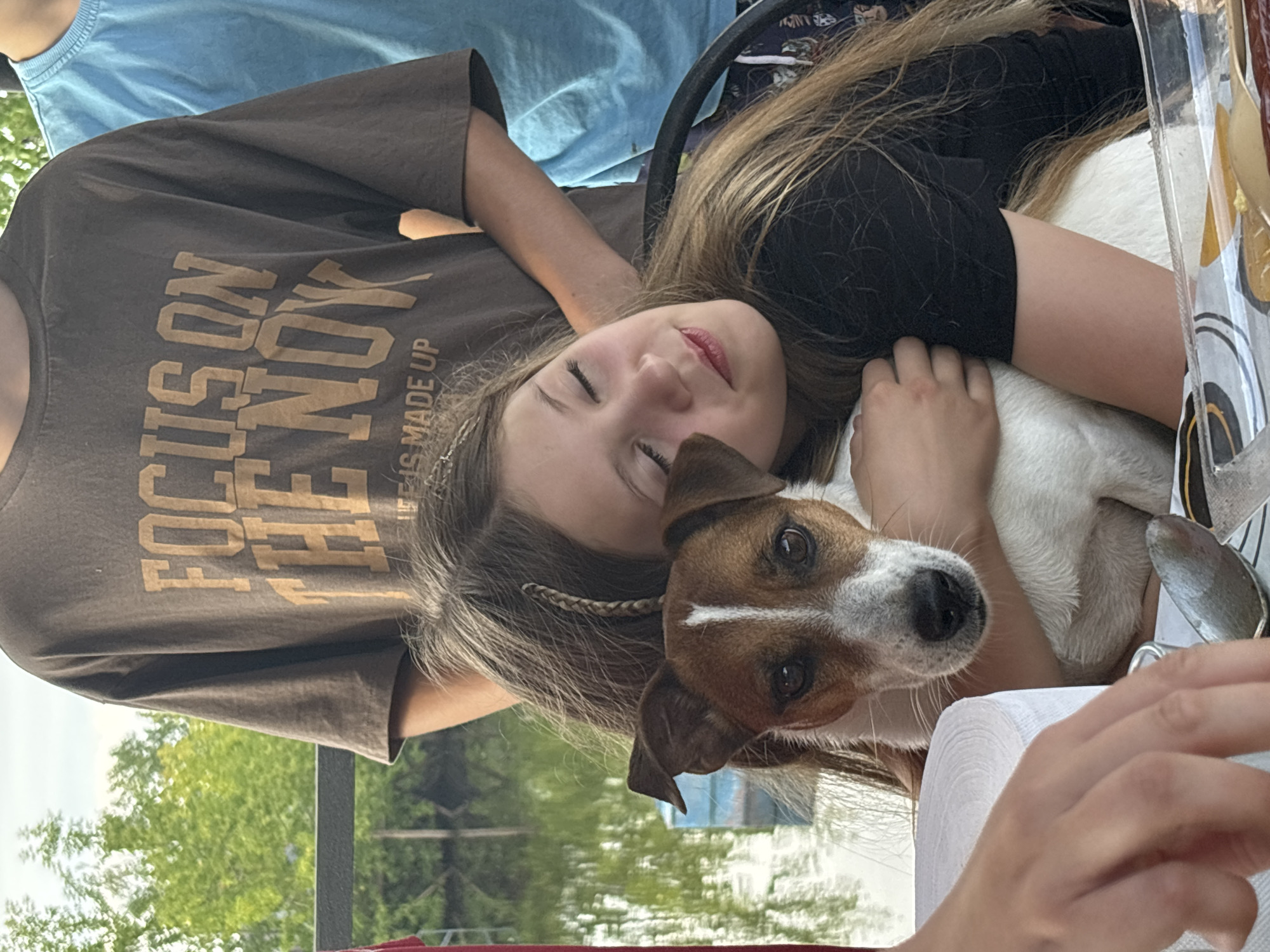 The image depicts a close-up of a young girl embracing a Jack Russell Terrier. The girl has long, blonde hair and her eyes are closed. The dog has a white and brown coat with distinct markings on its face. They appear to be outdoors, with a body of water and trees visible in the background. A person wearing a brown t-shirt with text on it, and another person in a blue shirt are also partially visible. The overall scene suggests a casual, relaxed moment. The image does not provide any explicit information about its location in the city of Chaikovsky, Russia, nor any details about specific forensic evidence.