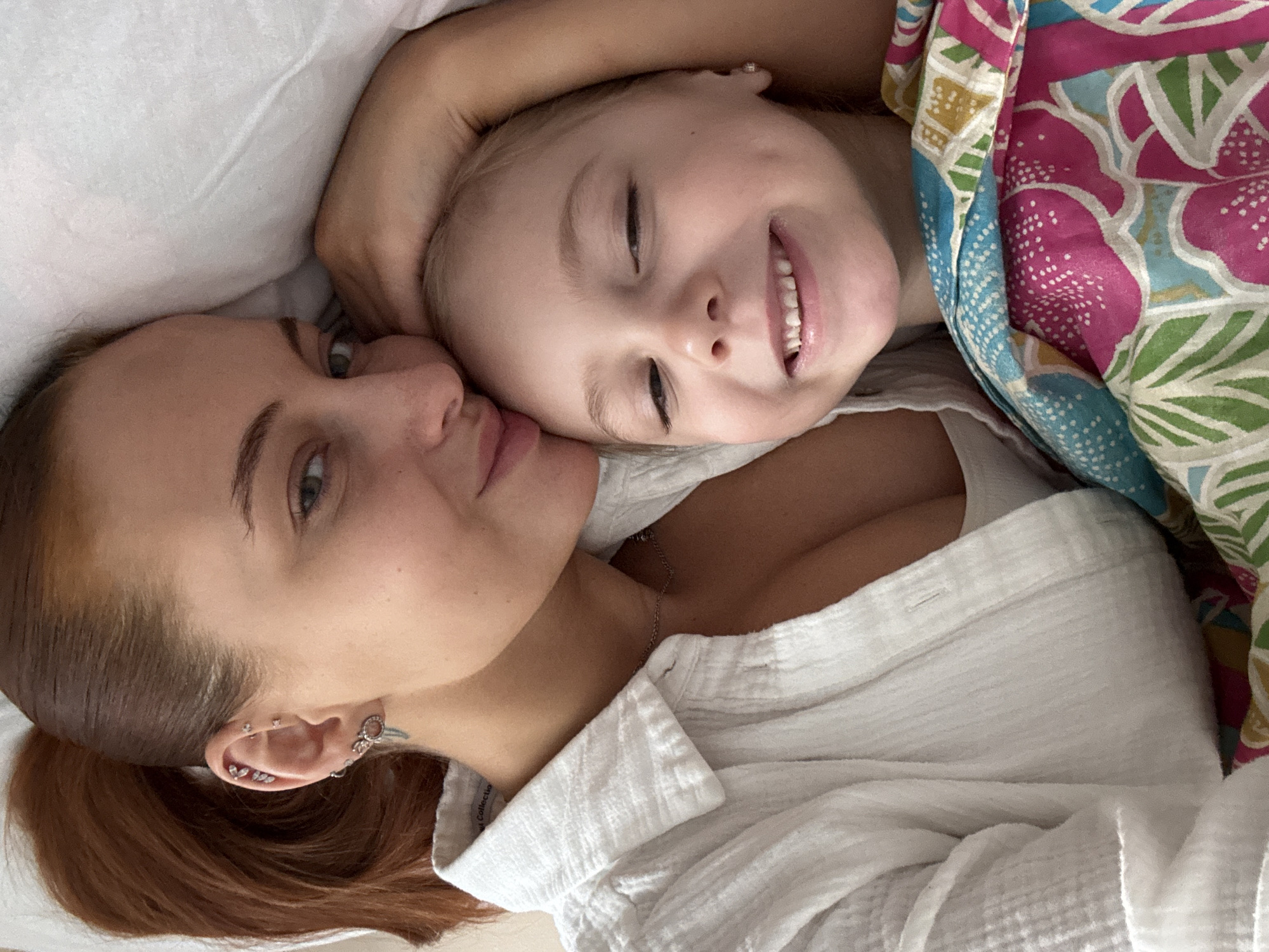 The image shows a woman and a young child lying in bed. The woman has auburn hair pulled back in a ponytail, fair skin, and multiple piercings in her left ear. She is wearing a white shirt and a silver necklace. The child has fair skin, blonde hair, and is smiling with closed eyes. They are under a patterned blanket with pink, green, blue, and yellow elements. The setting is indoors, presumably a bedroom. The city of Chastyye, Russia, cannot be confirmed from the visual evidence.