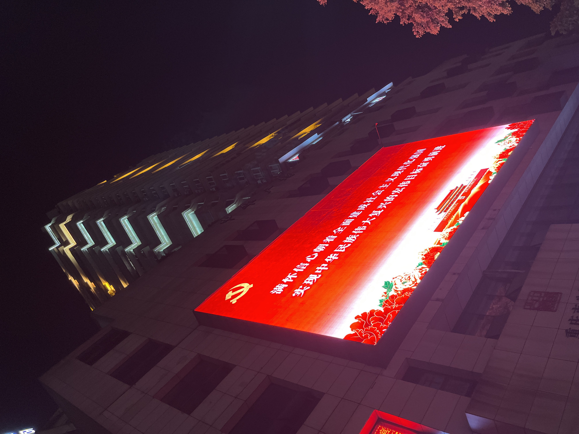 An exterior, low-angle, night shot captures a modern building in Yuzhong District, Chongqing, China. The building is illuminated with bright lights, highlighting its architecture. A large, red LED screen is prominently displayed on the side of the building, featuring Chinese characters and the symbol of the Communist Party of China. Below the LED screen, what appears to be an emblem is visible on the building's facade. To the right, the branches of a tree are partially visible against the dark night sky. The overall scene suggests a public space in an urban environment at night, with a focus on the illuminated building and its prominent display.