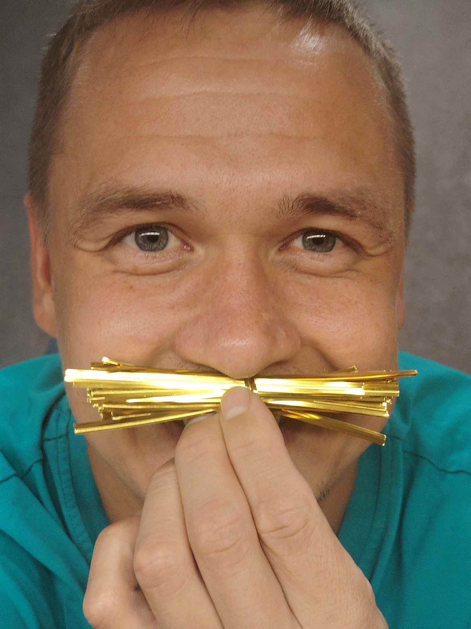 A man is holding a bunch of thin, golden metallic strands in front of his face, simulating a mustache. He is wearing a teal-colored shirt or uniform. The background is a plain, grayish surface. The man's face is visible, with his eyes looking directly at the camera. The image is a close-up shot, focusing on the man's face and the metallic strands. The specific location in Chaikovsky, Russia is not discernible from the provided image.