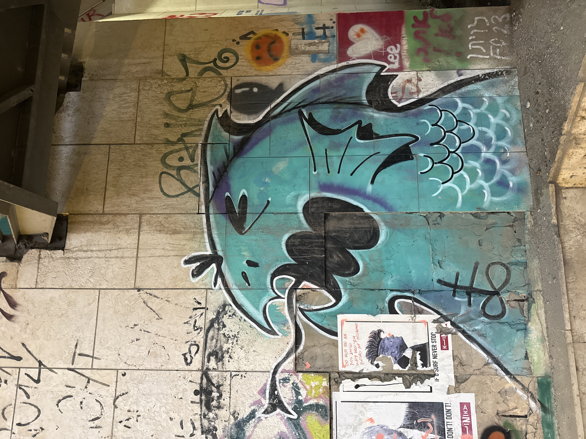 The image shows a wall covered in graffiti, likely in an urban outdoor setting in Tel Aviv, Israel, given the Hebrew text. The main subject is a large, stylized turquoise and purple fish painted on a tiled surface. The fish has a cartoonish, somewhat menacing expression with sharp fins and a long, trailing line from its mouth.
Around the fish, various other graffiti tags and drawings are visible. To the upper left, there's a grey metal structure, possibly part of a staircase or overhead covering. Above the fish, several smaller spray-painted elements include a yellow circle with a sad face, a stylized red heart with eyes and a ghost-like figure, and some Hebrew lettering. To the right of the fish, there are two posters adhered to the wall. The top poster features a drawing of a person with purple skin and spiky hair, accompanied by English text that reads "DO NOT BE AN ASS HOLE WHY ARE YOU BEING AN ASSHUE" and "IF SURF NEVER STOP". Below this, another poster has red lettering with the words "DON'T! DON'T!" and "LINIK".
Further Hebrew text, "לויתן" (Leviathan), is visible on the wall, and some indistinct black spray paint tags are scattered across the tiled sections. The overall impression is one of urban decay and street art, common in alleyways or underpasses. The lighting suggests it might be daytime, but the environment appears to be somewhat shaded.